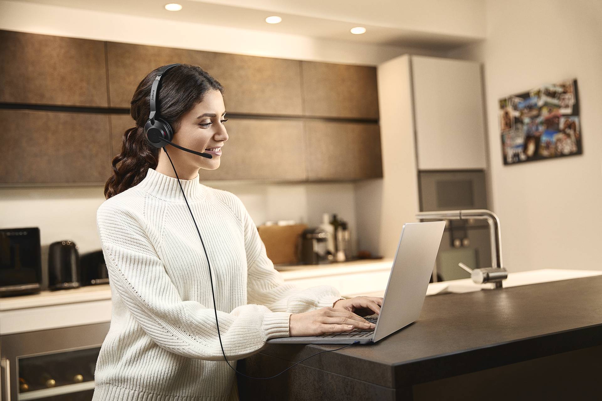 Frau mit Headset steht in moderner Küche und tippt auf Laptop. Sie sieht fokussiert aus, trägt weißen Pullover.