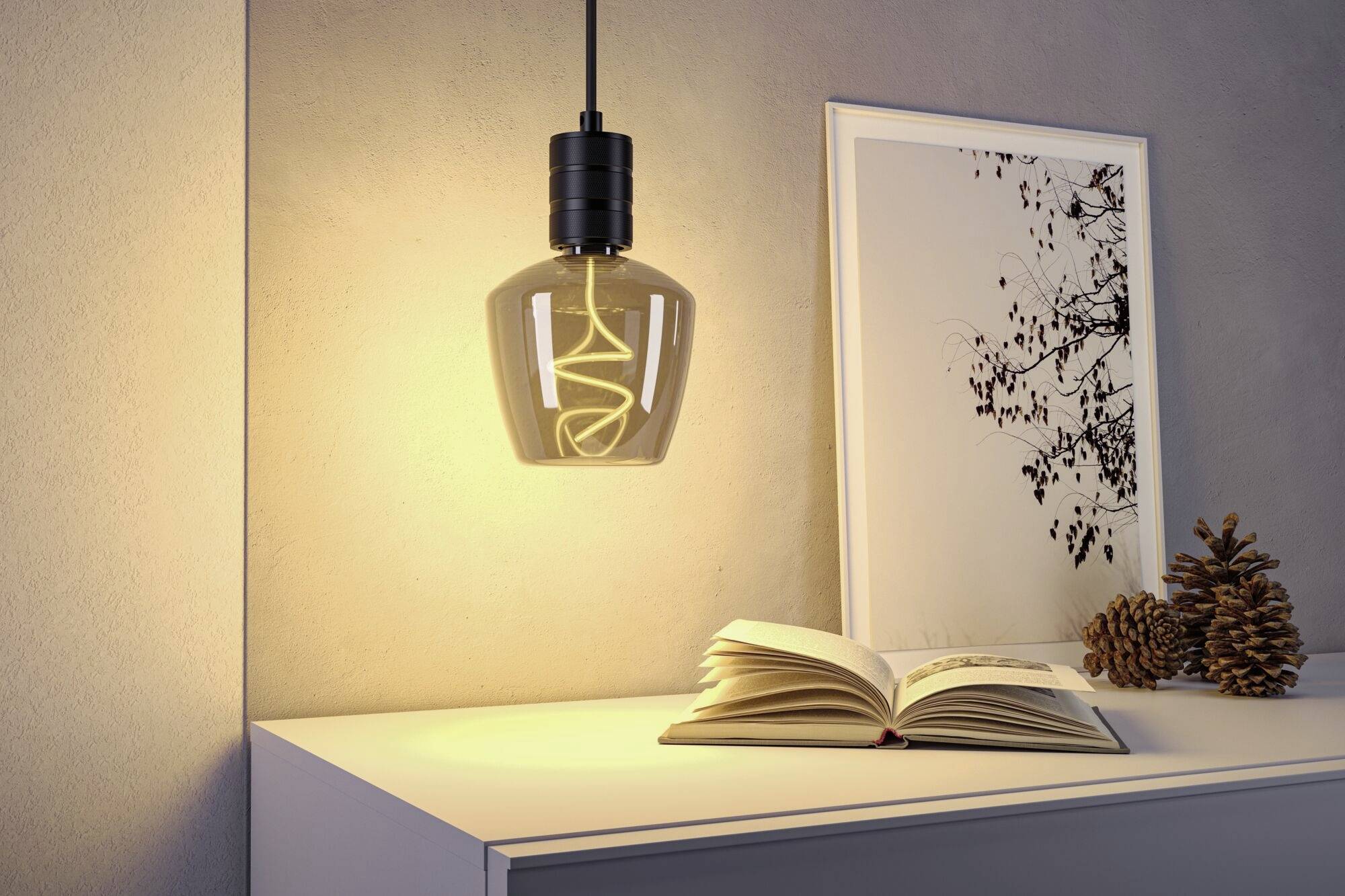 Eine stilvolle Lampe leuchtet über einem Tisch mit einem offenen Buch, daneben steht ein gerahmtes Kunstbild und einige Kiefernzapfen.