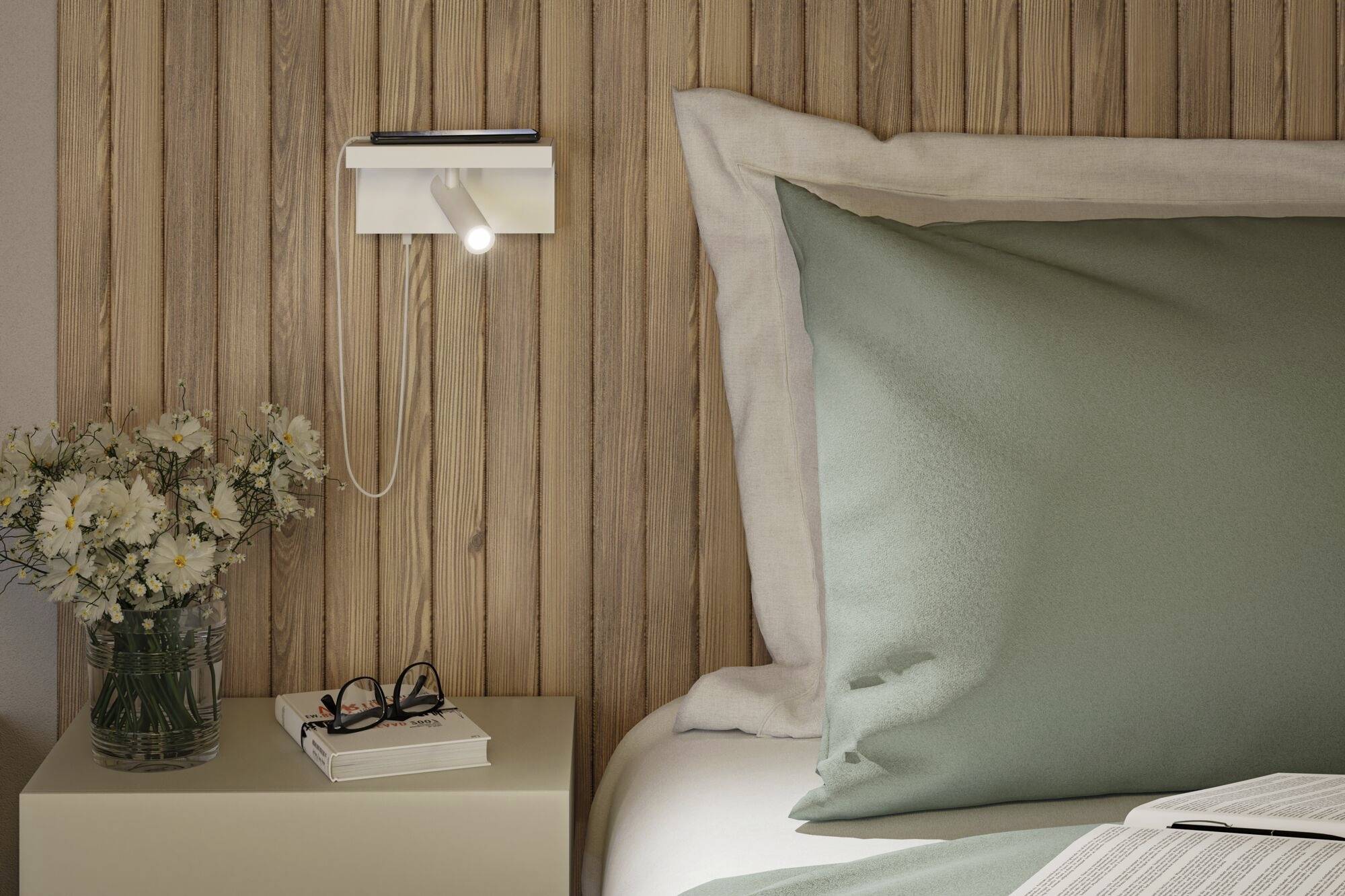 Ein gemütliches Schlafzimmer mit einem Bett, grünen Kissen und einer Holzwand. Nachttisch mit Leselampe, Buch, Brille und Blumen.