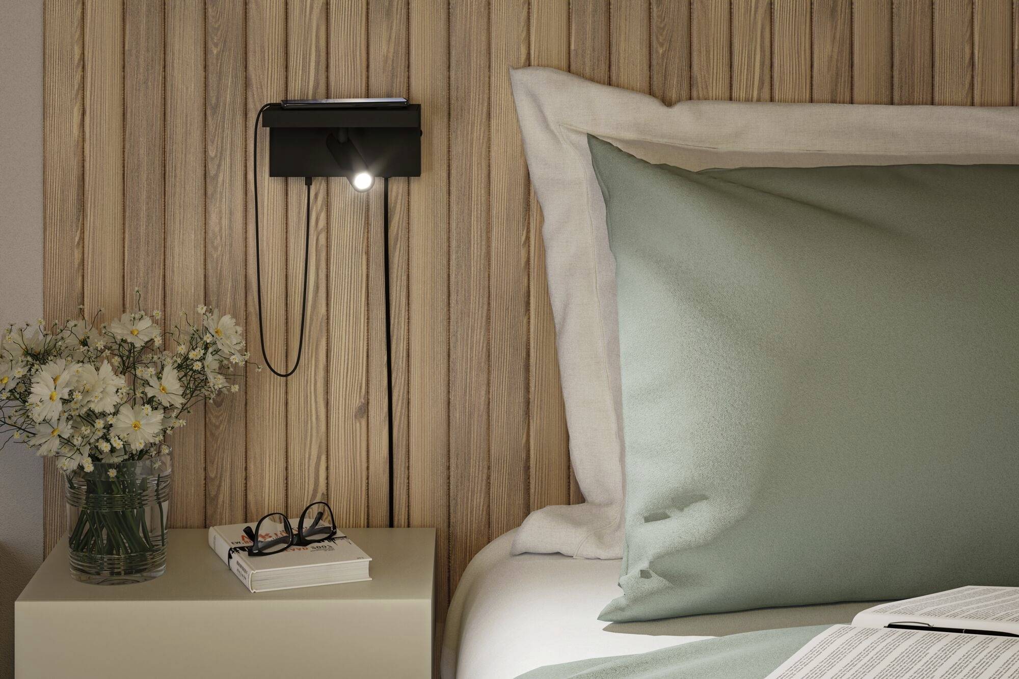 Schlafzimmer mit Holzrückwand, einem Nachttisch, darauf eine Vase mit Blumen, Bücher, eine Brille und eine kleine Wandleuchte.