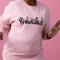 Eine Person trägt einen rosa Pullover mit der Aufschrift 'Grandma' und Blumenmotiven darauf, vor einem rosa Hintergrund.