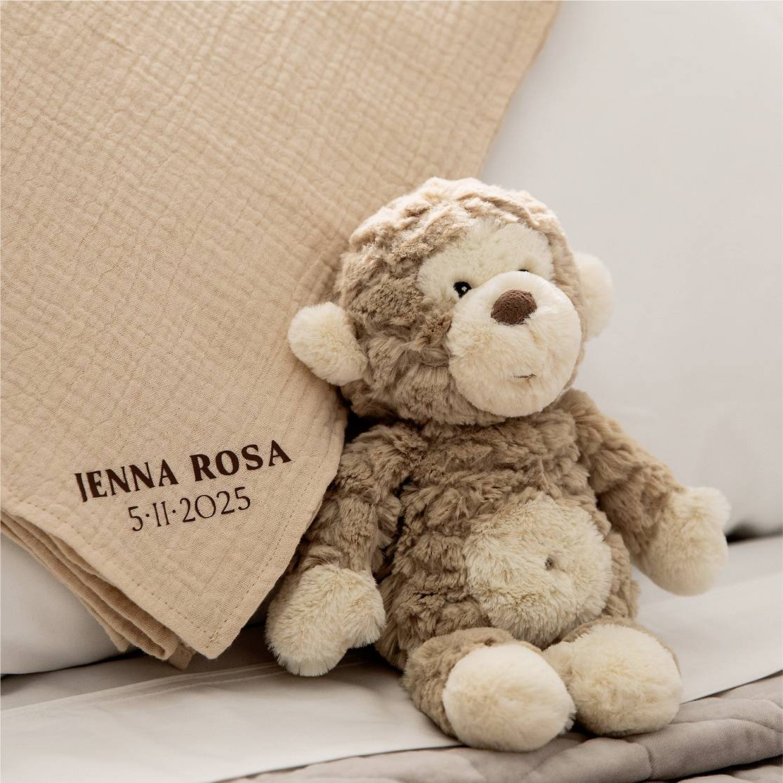 Ein brauner Plüschaffe sitzt auf einem Bett neben einer beigen Decke, auf der 'Jenna Rosa 5-11-2025' gestickt ist.