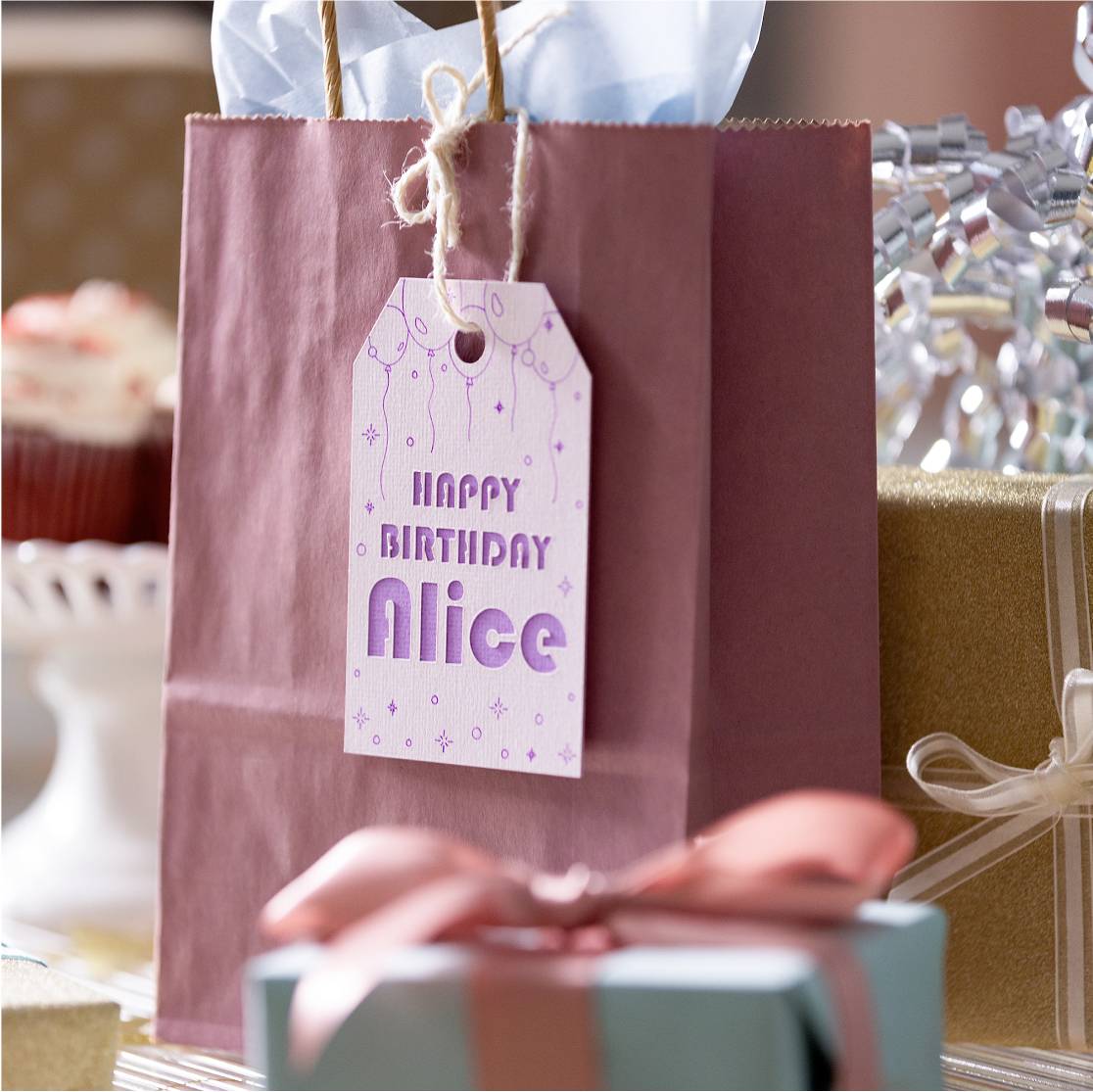 'Happy Birthday Alice' steht auf einer Geschenktüte, daneben ein Geschenk in rosafarbener Schleife. Hintergrund unscharf mit Partydekoration.