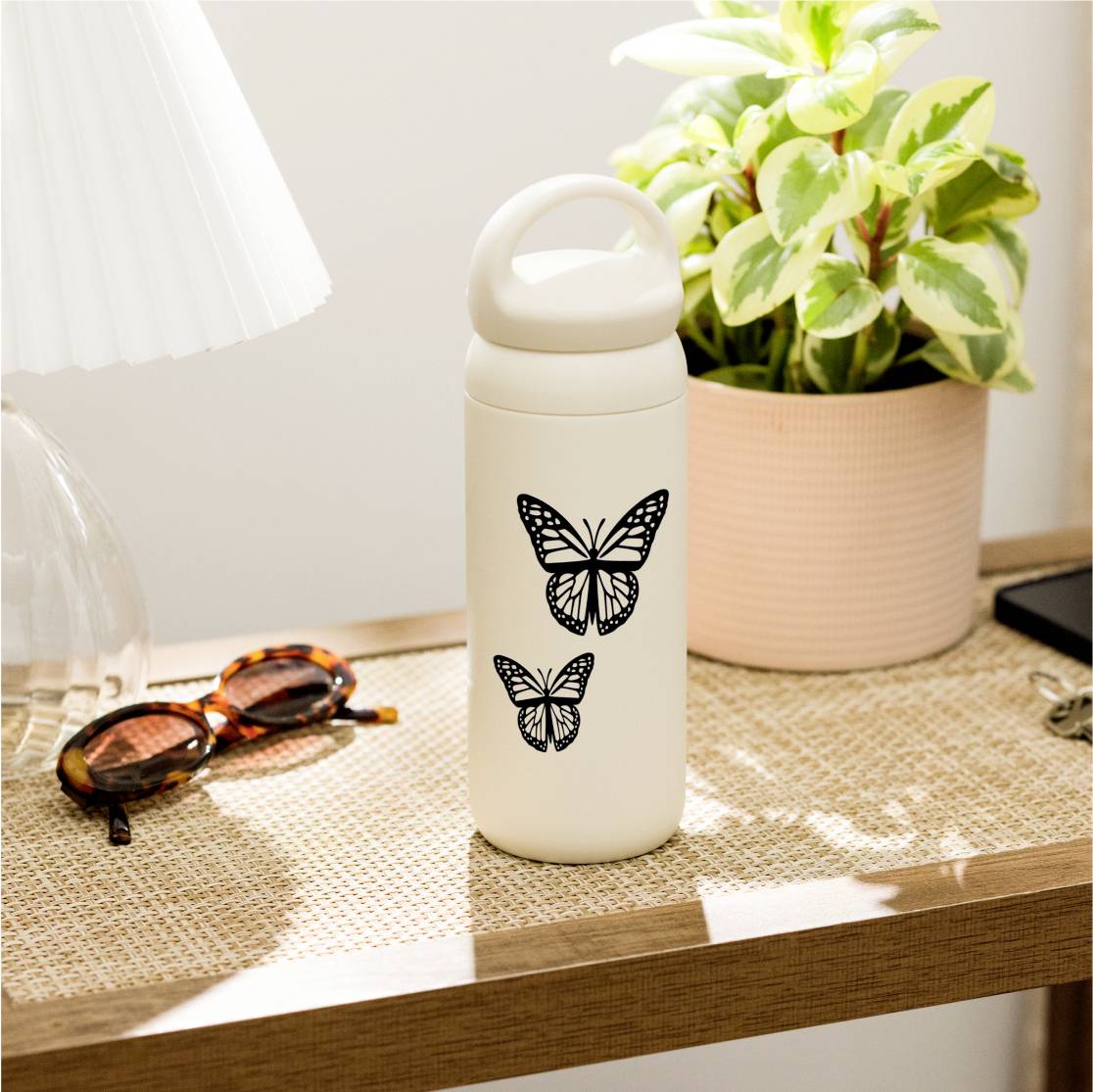 Eine weiße Wasserflasche mit schwarzen Schmetterlingsmotiven steht auf einem Tisch neben einer Topfpflanze und einer Sonnenbrille.