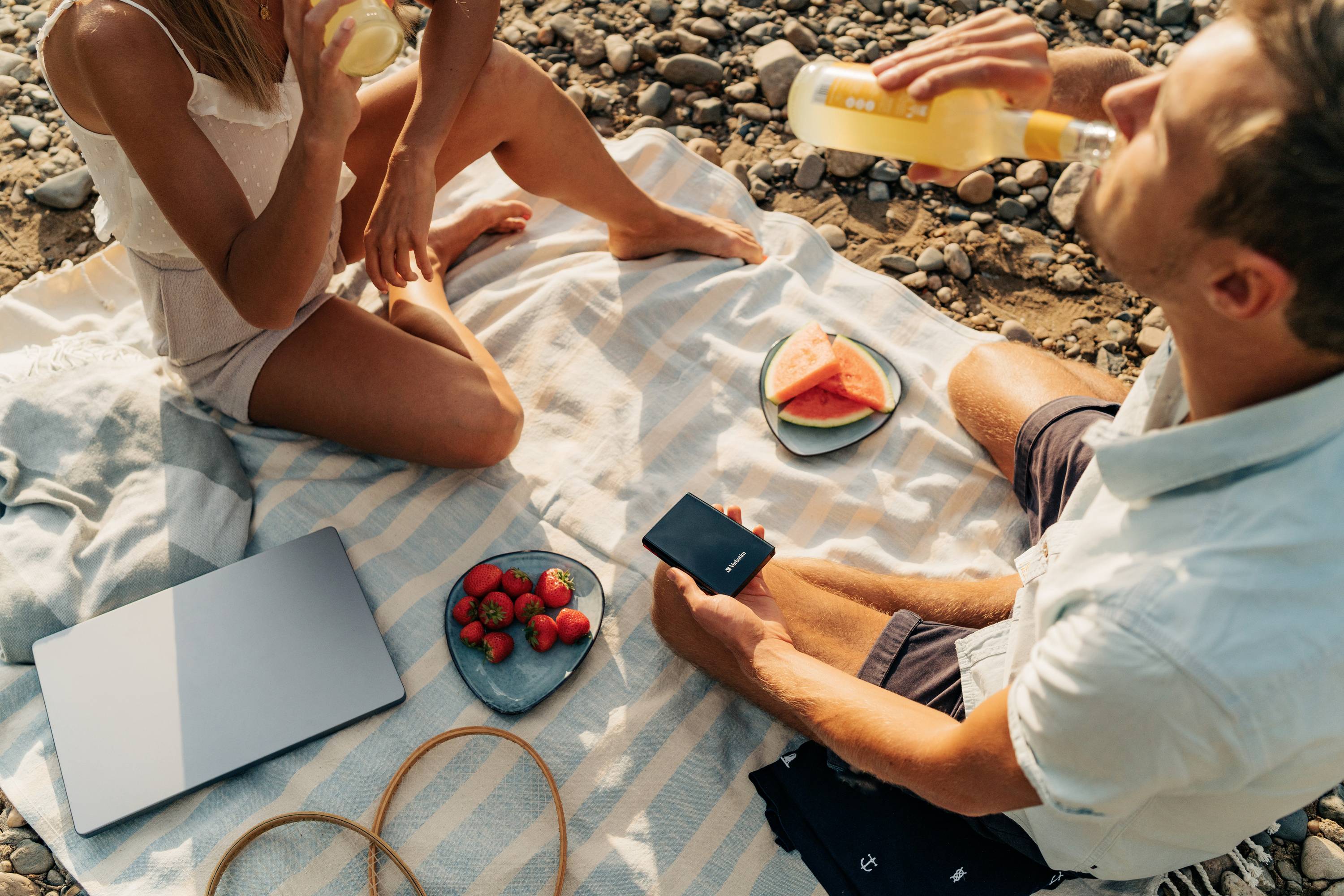 Ein Paar sitzt auf einer Picknickdecke am Strand, isst Wassermelone und Erdbeeren und trinkt aus Flaschen. Ein Laptop liegt daneben.