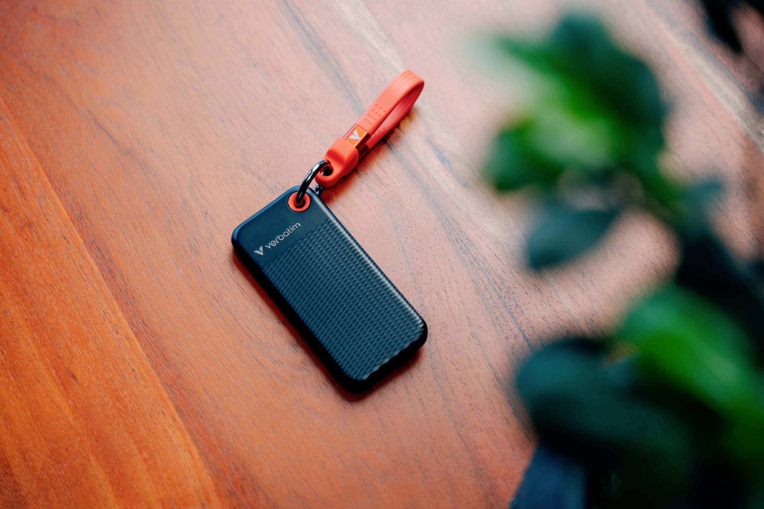 Eine schwarze tragbare Festplatte mit rotem Band liegt auf einem Holztisch. Im Vordergrund ist unscharf eine Pflanze zu sehen.