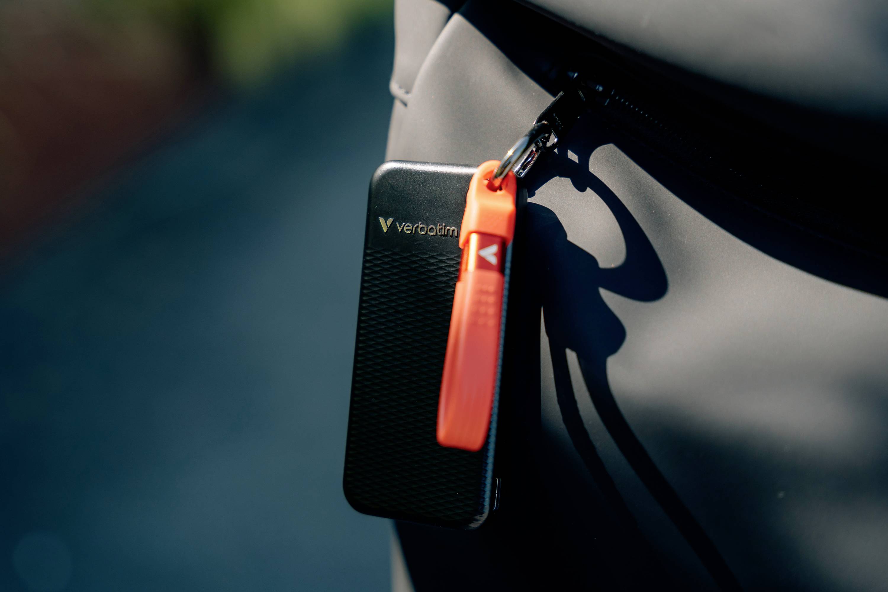 Eine schwarze externe Festplatte der Marke Verbatim mit einem orangefarbenen Schlüsselanhänger, die an einem Reißverschluss befestigt ist.