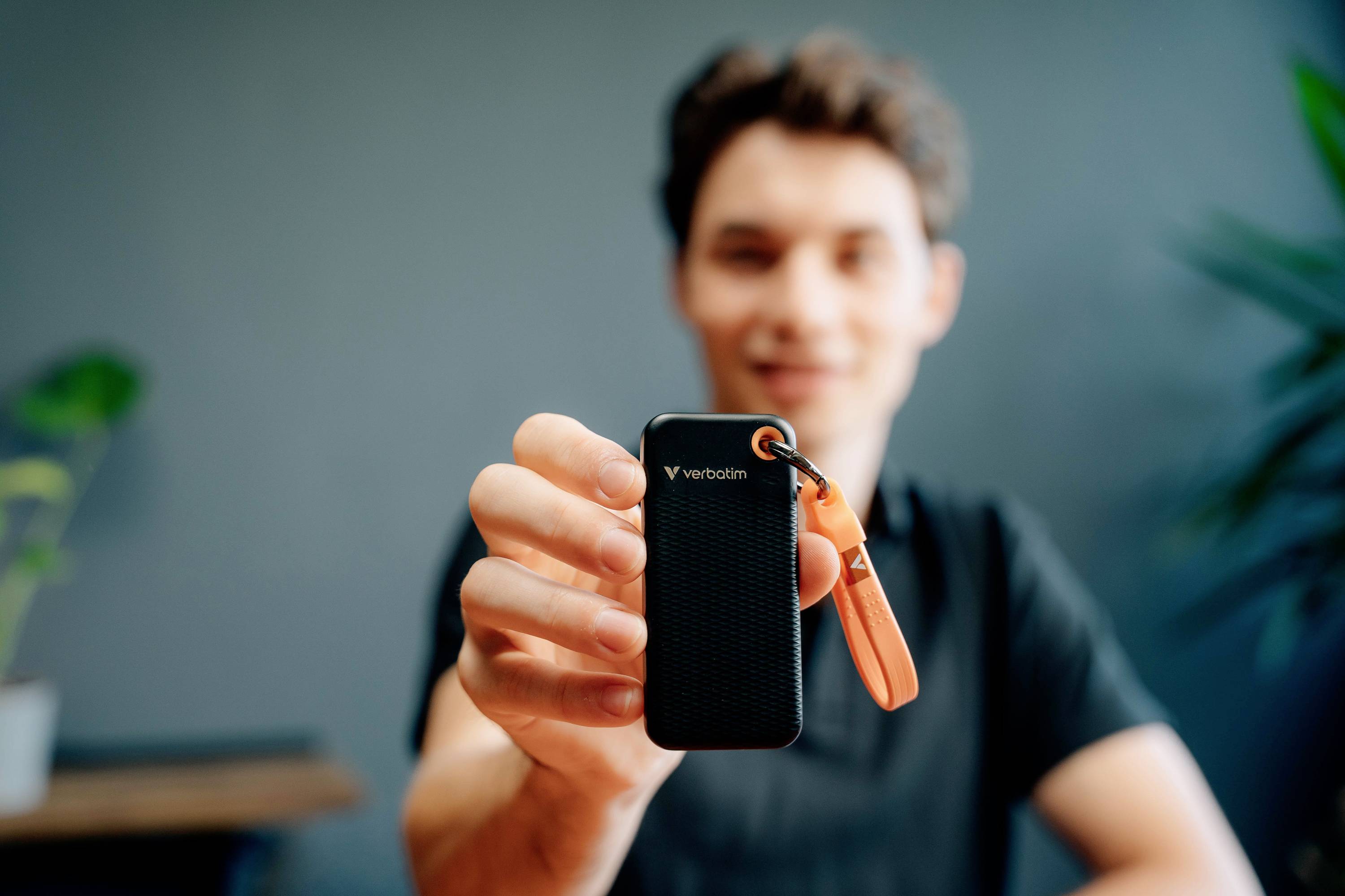 Eine Person hält eine tragbare externe Festplatte mit einem orangefarbenen Anhänger in die Kamera. Im Hintergrund Pflanzen und unscharfe Möbel.