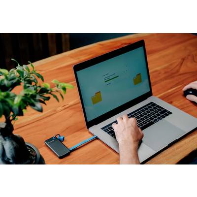 Eine Person benutzt einen Laptop auf einem Tisch. Ein externer Speicher ist mit Kabel verbunden, während ein Dateiordner auf dem Bildschirm angezeigt wird.