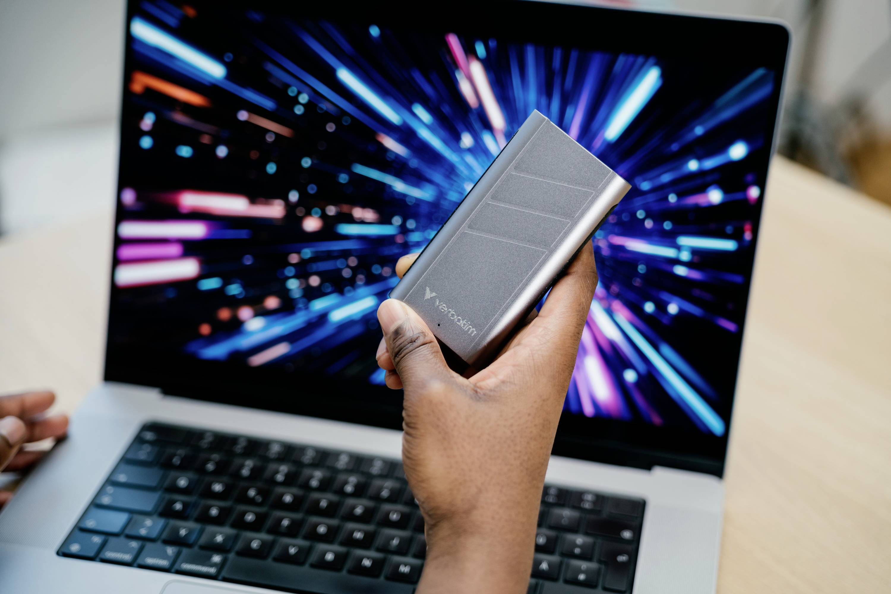Hand hält eine externe Festplatte vor einem Laptop, auf dessen Bildschirm farbige Lichter in Bewegung dargestellt sind.
