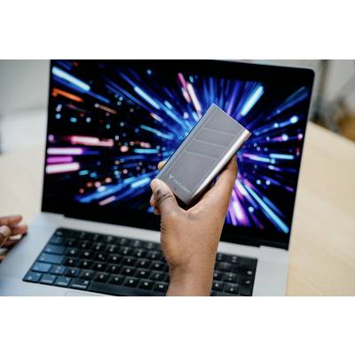 Hand hält eine externe Festplatte vor einem Laptop, auf dessen Bildschirm farbige Lichter in Bewegung dargestellt sind.