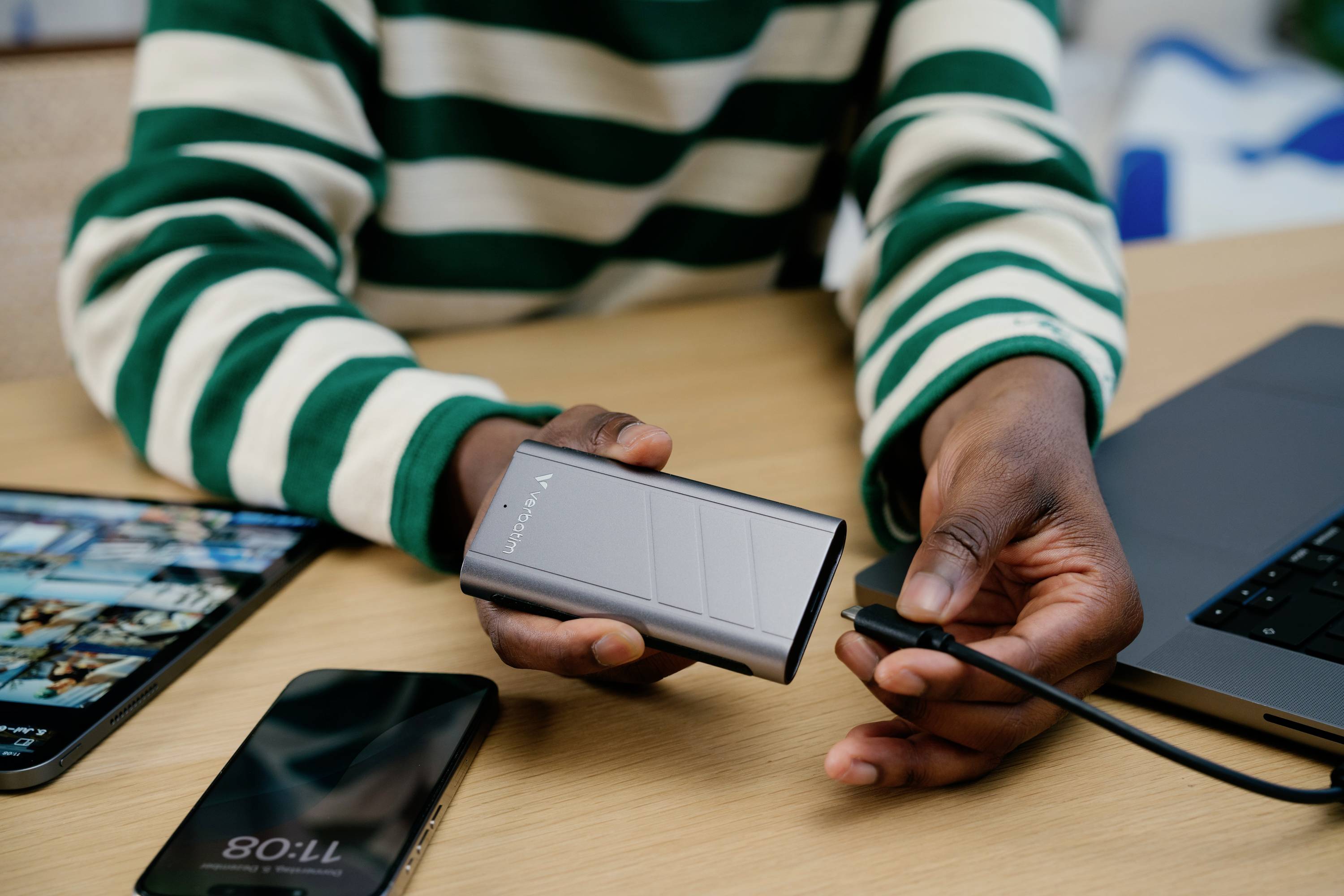 Eine Person hält eine Powerbank und ein Ladekabel, bereit zum Anschluss. Im Hintergrund liegt ein Laptop und ein Smartphone auf dem Tisch.