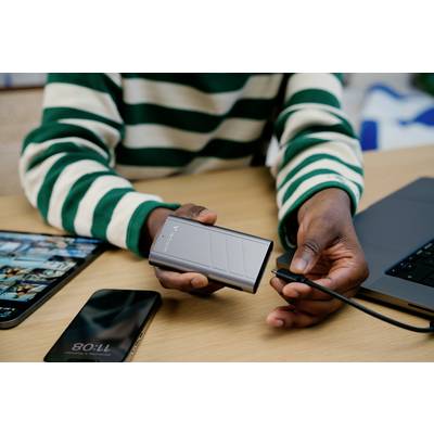 Eine Person hält eine Powerbank und ein Ladekabel, bereit zum Anschluss. Im Hintergrund liegt ein Laptop und ein Smartphone auf dem Tisch.