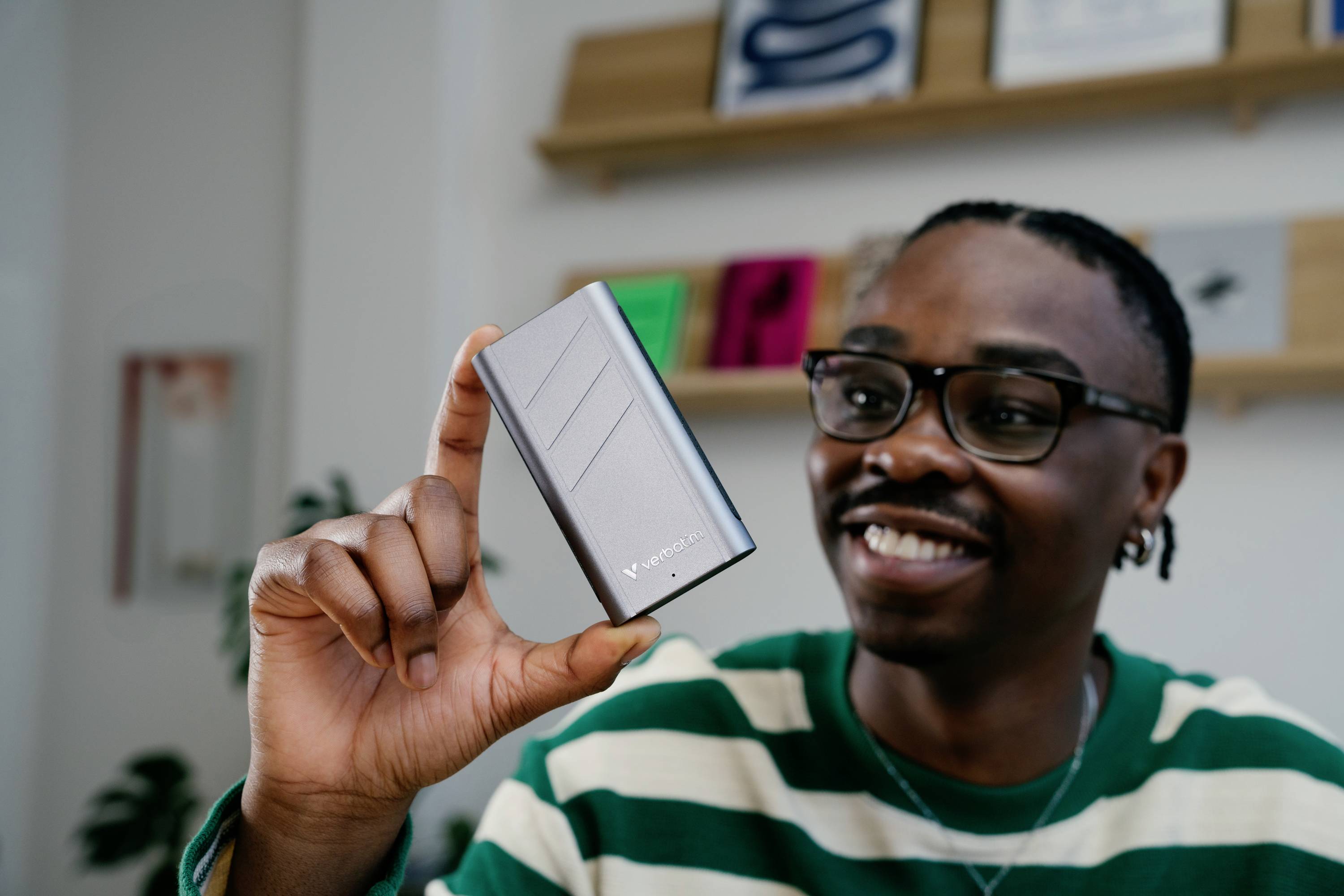 Eine Person hält eine tragbare Festplatte, lächelnd. Im Hintergrund sind Bücher und Rahmen auf einem Regal sichtbar.