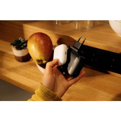 Eine Hand hält ein weißes und ein schwarzes Ladegerät neben einem Tisch mit einem Apfel und einer Topfpflanze in einem gut beleuchteten Raum.