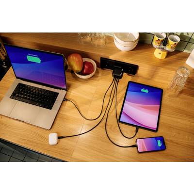 Ein Laptop, ein Tablet und ein Smartphone werden auf einem Holztisch über eine Mehrfachsteckdose aufgeladen, daneben Obstschale.