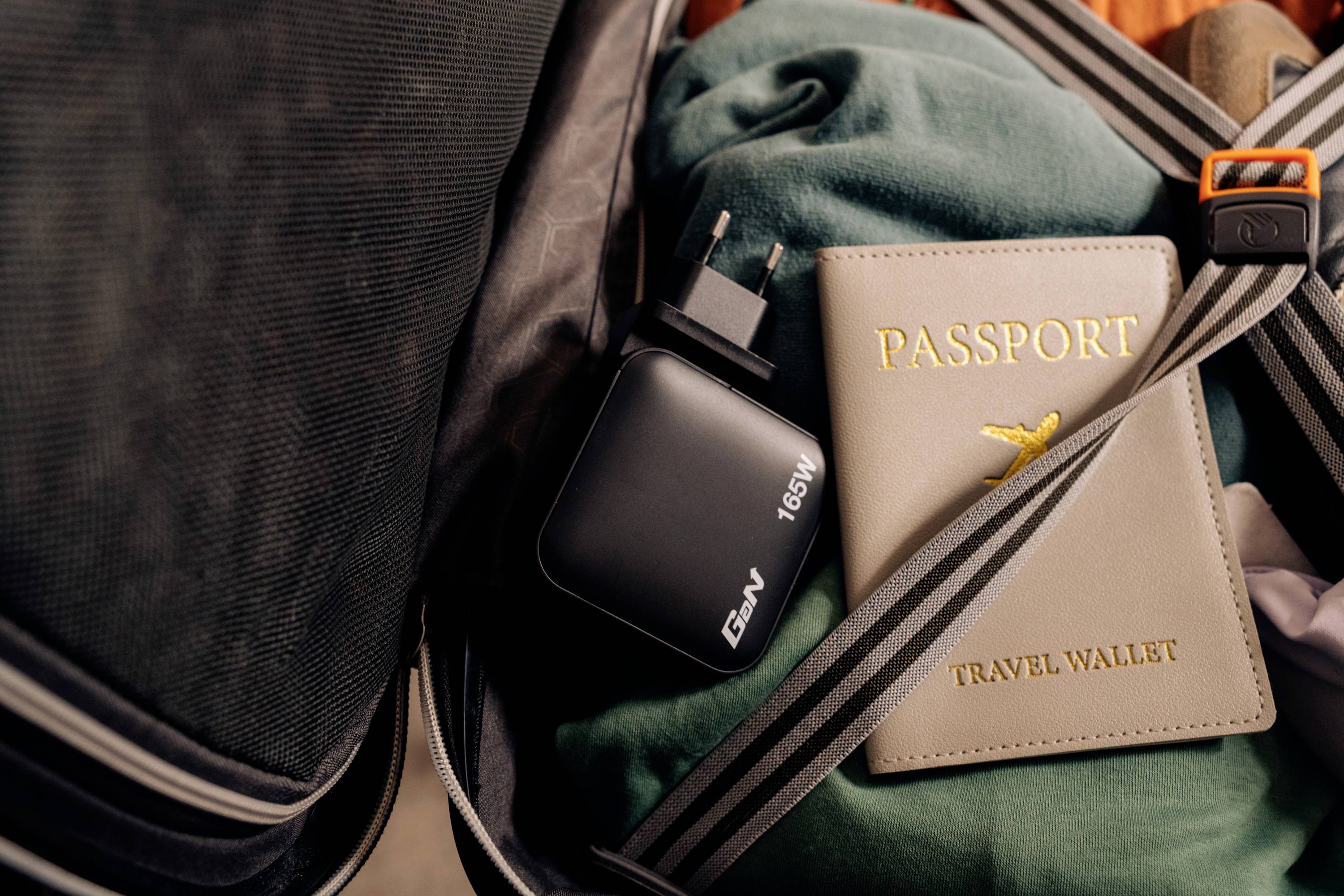Offener Koffer mit einem 65W-Ladegerät und einem 'Passport'-Reiseetui auf grüner Kleidung, gesichert mit einem Gurt.