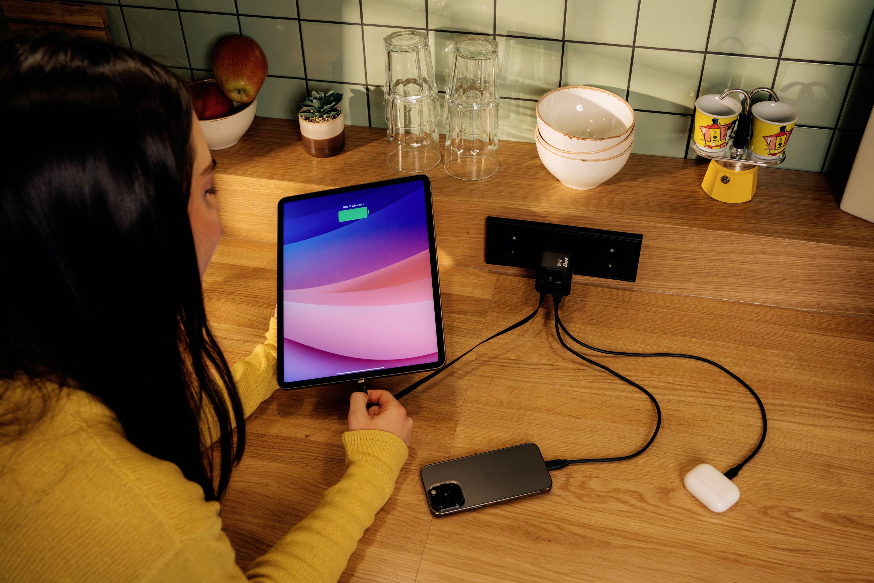 Eine Person lädt ein Tablet, ein Smartphone und Kopfhörer auf. Alle Geräte sind über Kabel mit einer Steckdosenleiste auf einem Tisch verbunden.