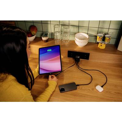 Eine Person lädt ein Tablet, ein Smartphone und Kopfhörer auf. Alle Geräte sind über Kabel mit einer Steckdosenleiste auf einem Tisch verbunden.