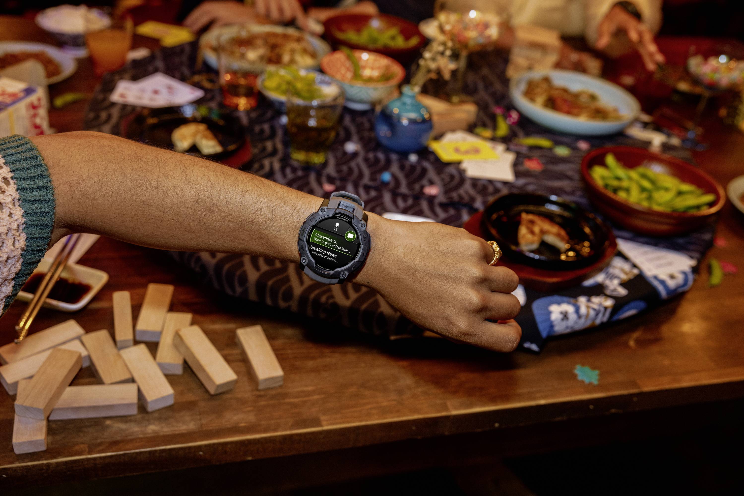 Eine Person zeigt eine Smartwatch an einem Handgelenk. Im Hintergrund ein Tisch mit Speisen, Getränken und einem Jenga-Spiel.