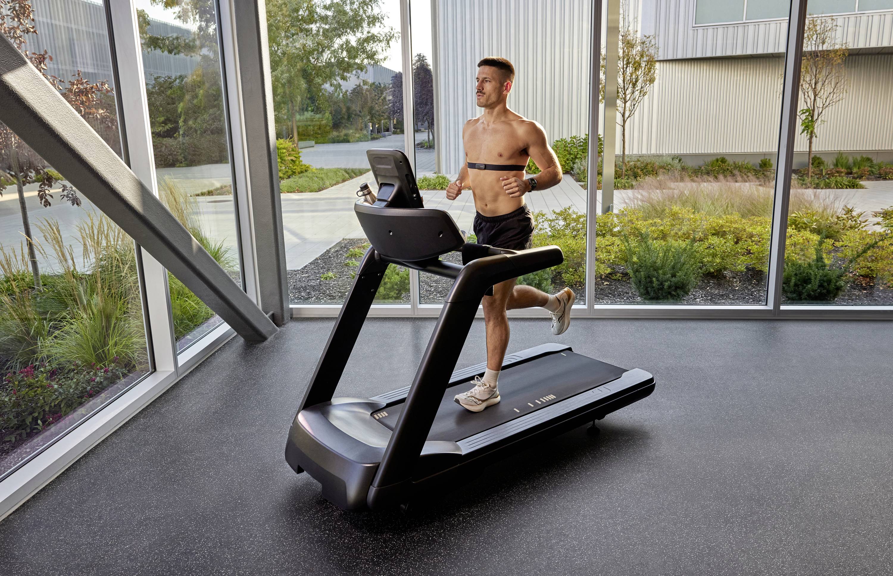 Ein Mann joggt auf einem Laufband in einem Fitnessstudio mit großen Fenstern, durch die man eine Gartenlandschaft sieht.