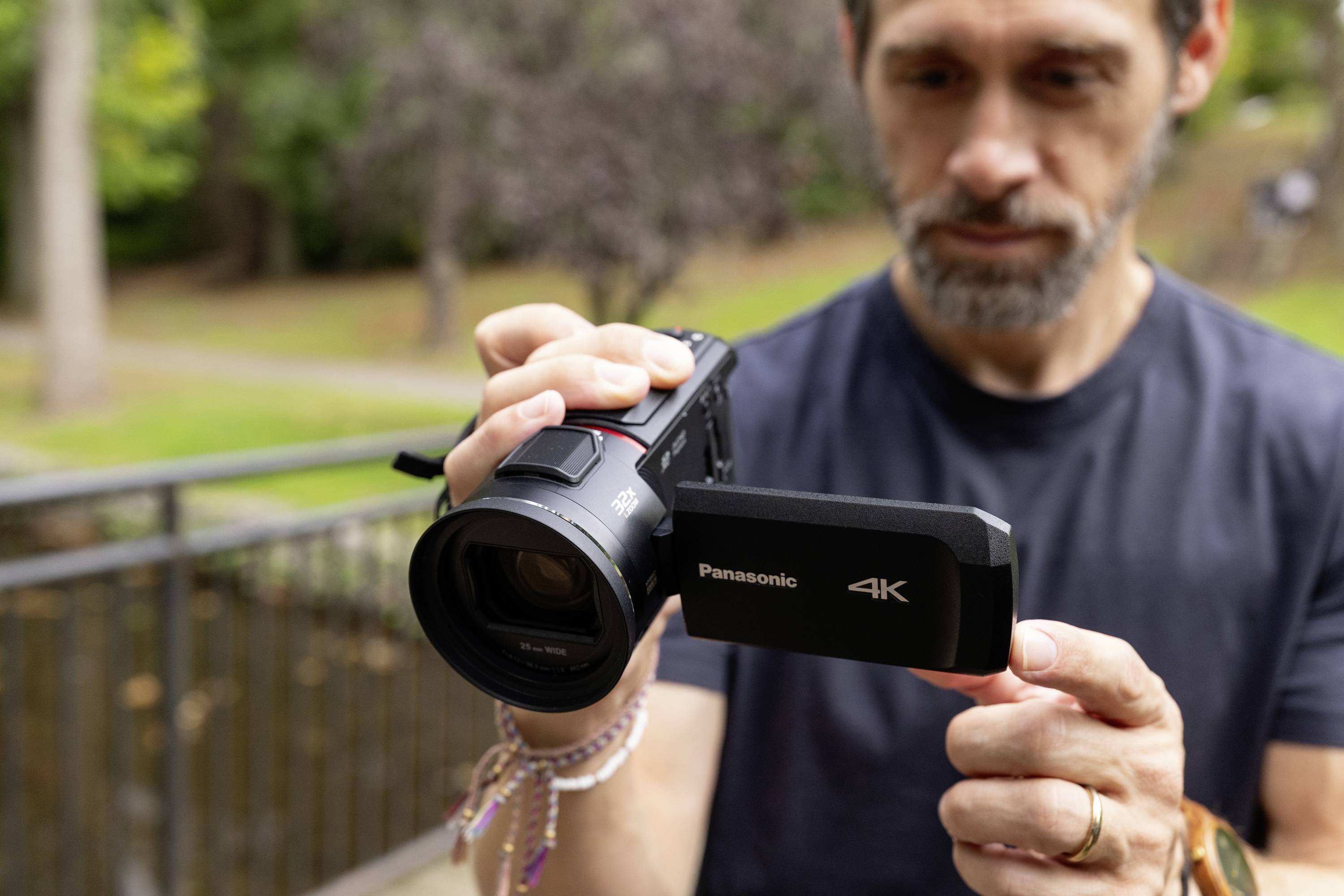 Ein Mann hält eine Panasonic 4K-Videokamera im Freien und überprüft vorsichtig das Objektiv. Im Hintergrund sind Bäume sichtbar.