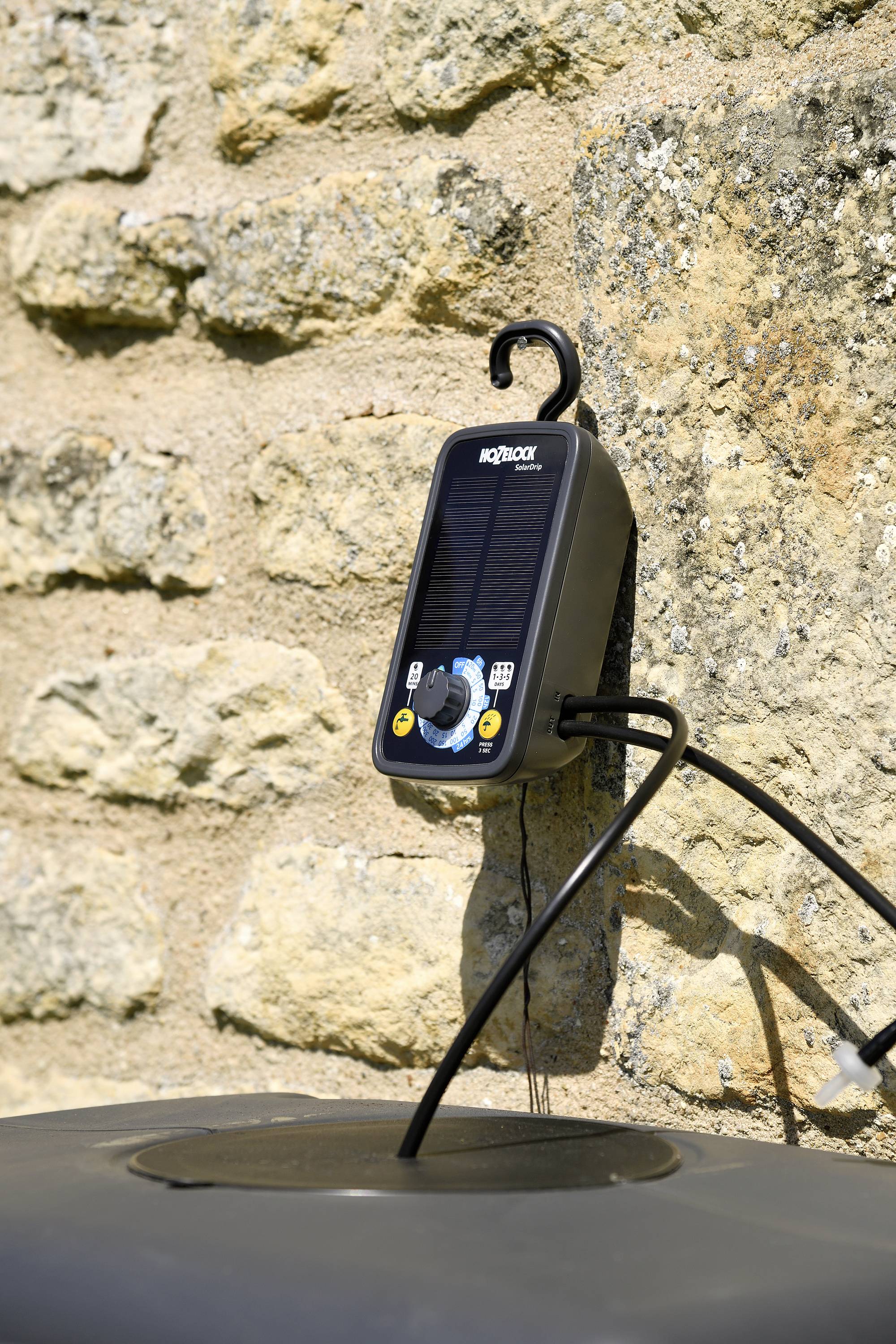 Solarbetriebene Wasserpumpensteuerung, montiert an einer Steinwand, verbunden mit einem Wasserschlauch. Ideal für Gartenbewässerung.
