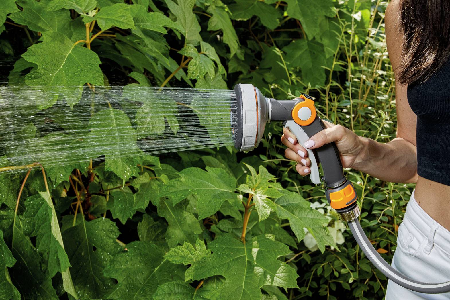 Eine Person gießt Pflanzen im Garten mit einem Gartenschlauch, aus dem Wasser in einem Sprühstrahl austritt. Sie hält den Schlauch mit der rechten Hand.