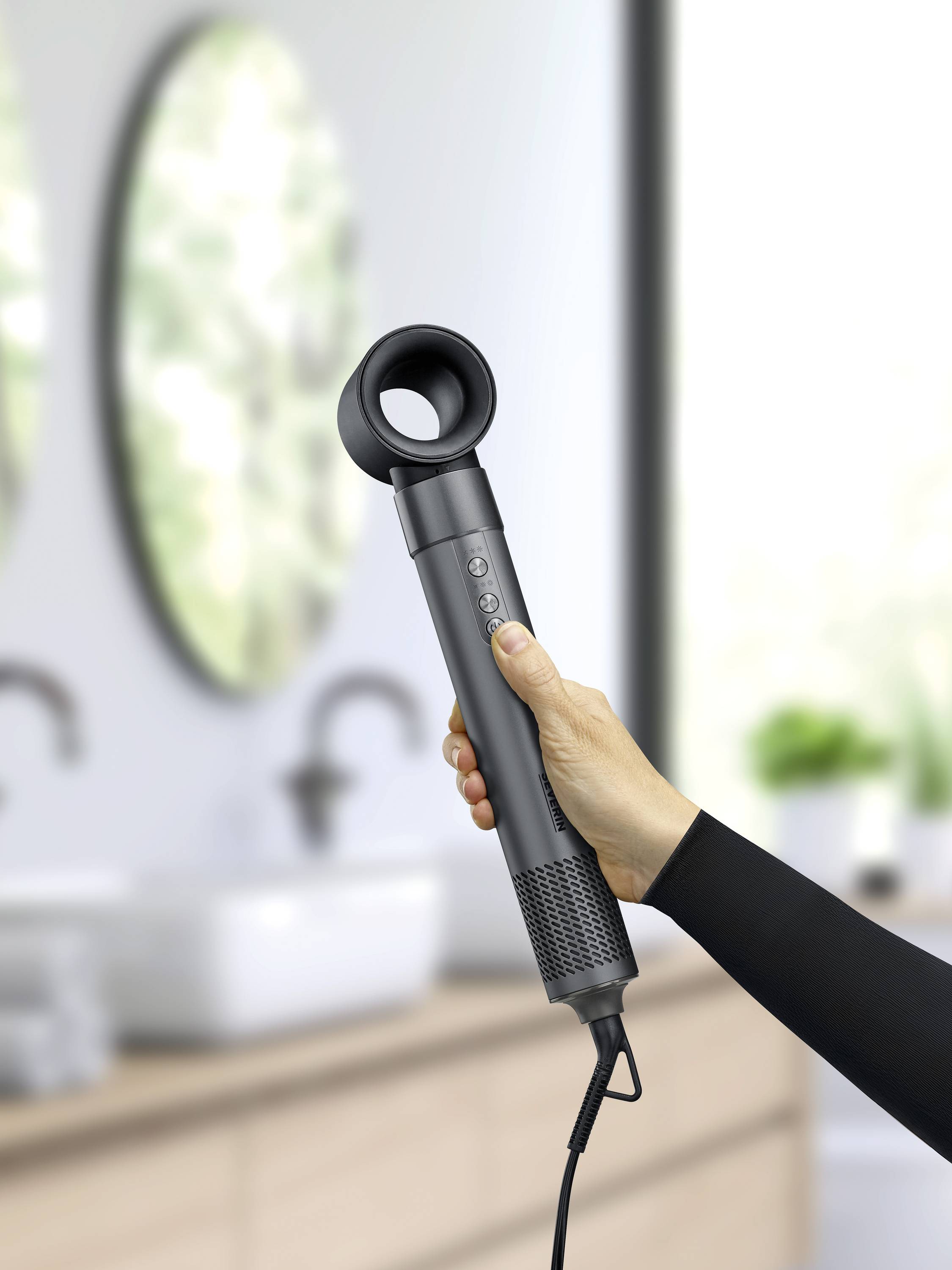 Eine Hand hält einen modernen, grauen Haartrockner in einem Badezimmer mit verschwommenem Hintergrund.