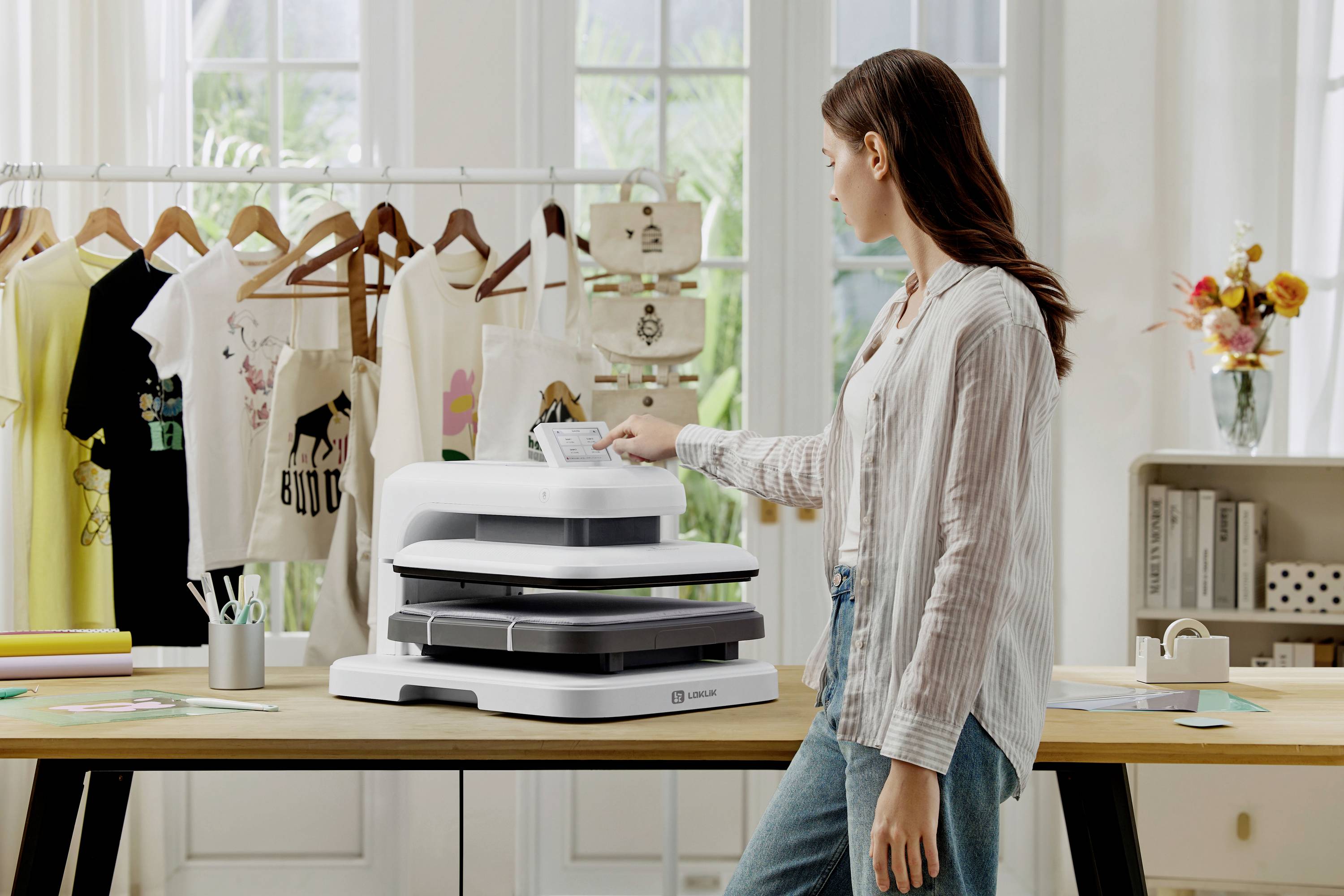 Eine Frau steht in einem Atelier vor einem Tisch mit Druckgerät, umgeben von T-Shirts an einem Kleiderständer und Bastelmaterialien.