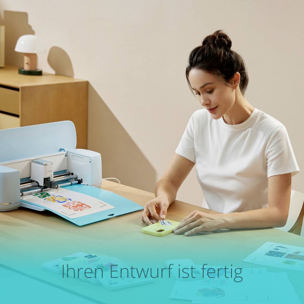 Eine Person in weißem T-Shirt arbeitet an einem Schneideplotter. Auf dem Bildschirm steht 'Ihr Entwurf ist fertig'.