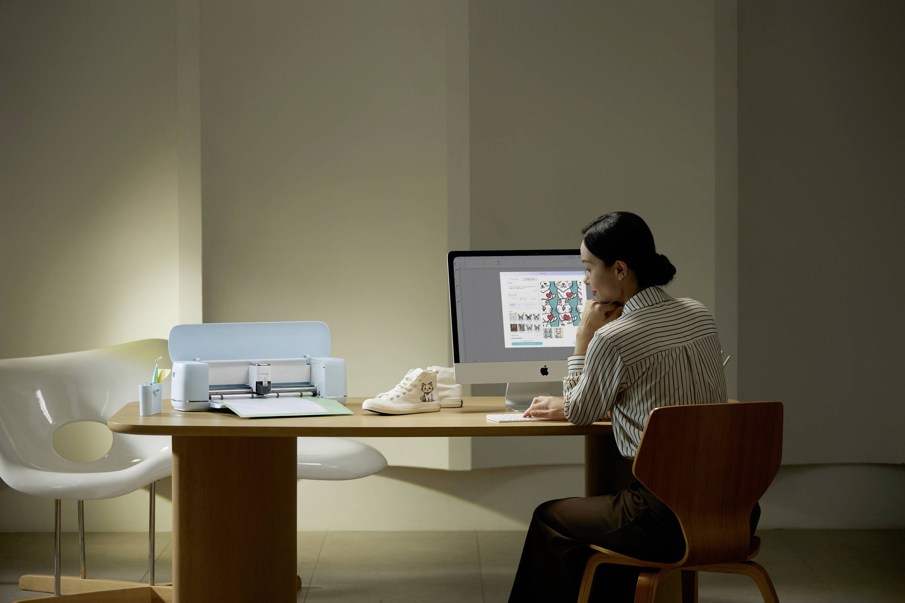 Eine Frau sitzt an einem Schreibtisch, betrachtet einen Computermonitor mit Grafikdesigns. Neben ihr steht ein Schneideplotter.