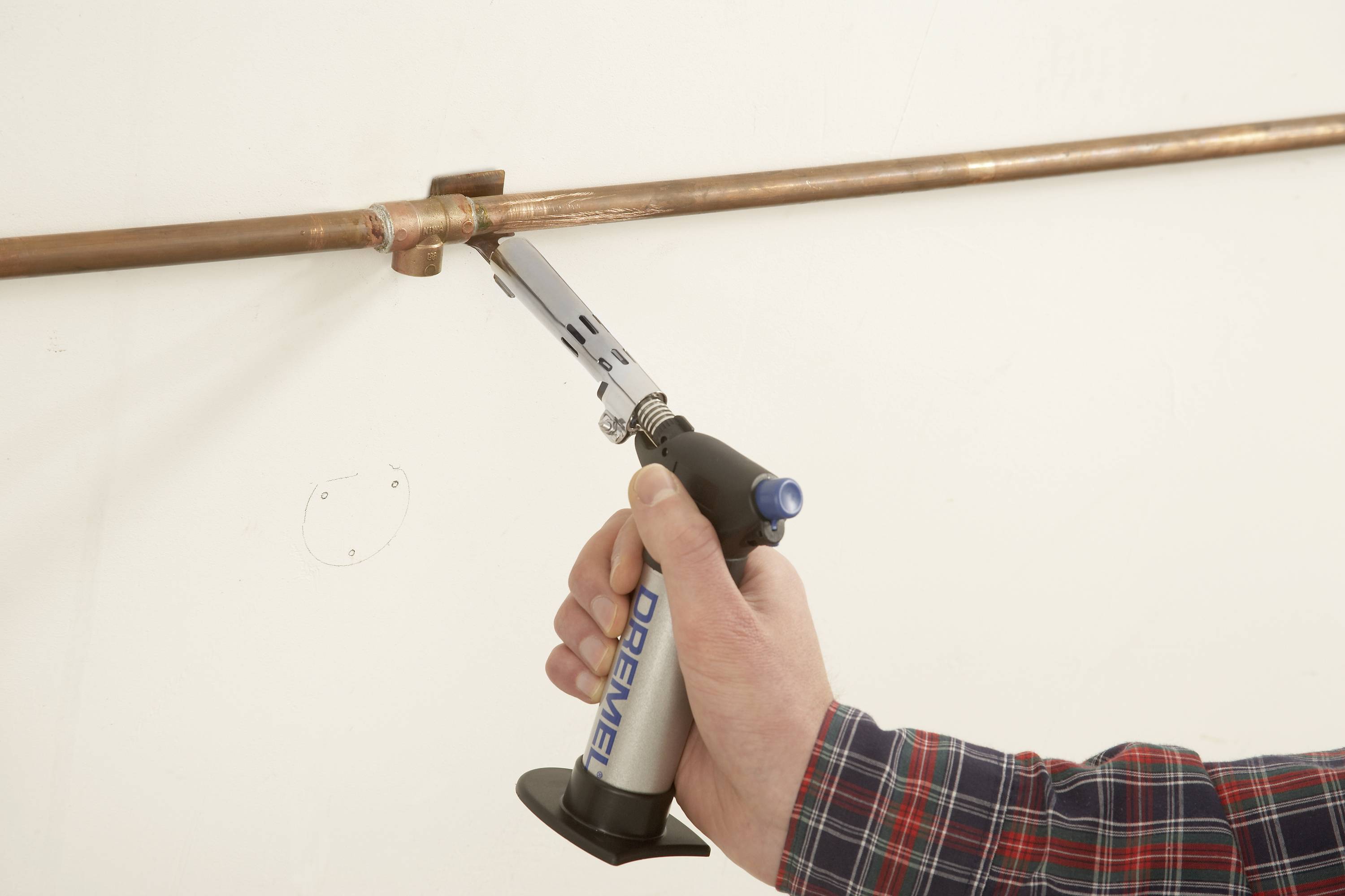 'Eine Person lötet ein Kupferrohr mit einem Handbrenner an einer weißen Wand. Der Brenner liegt in der rechten Hand.'