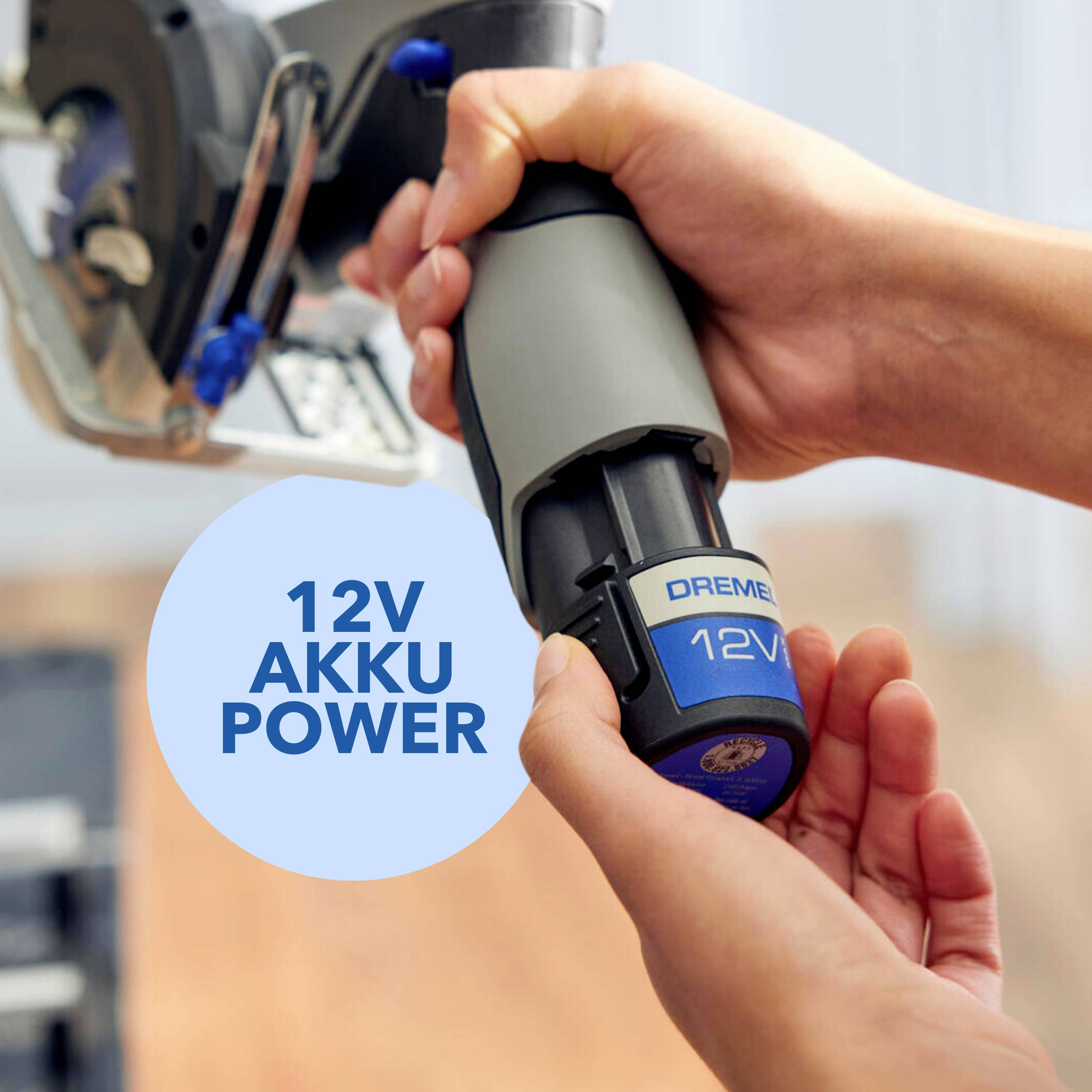 Person hält ein Dremel 12V Akku-Werkzeug. Im Hintergrund unscharfes Werkzeug sichtbar. Fokus auf Akkugerät in der Hand.