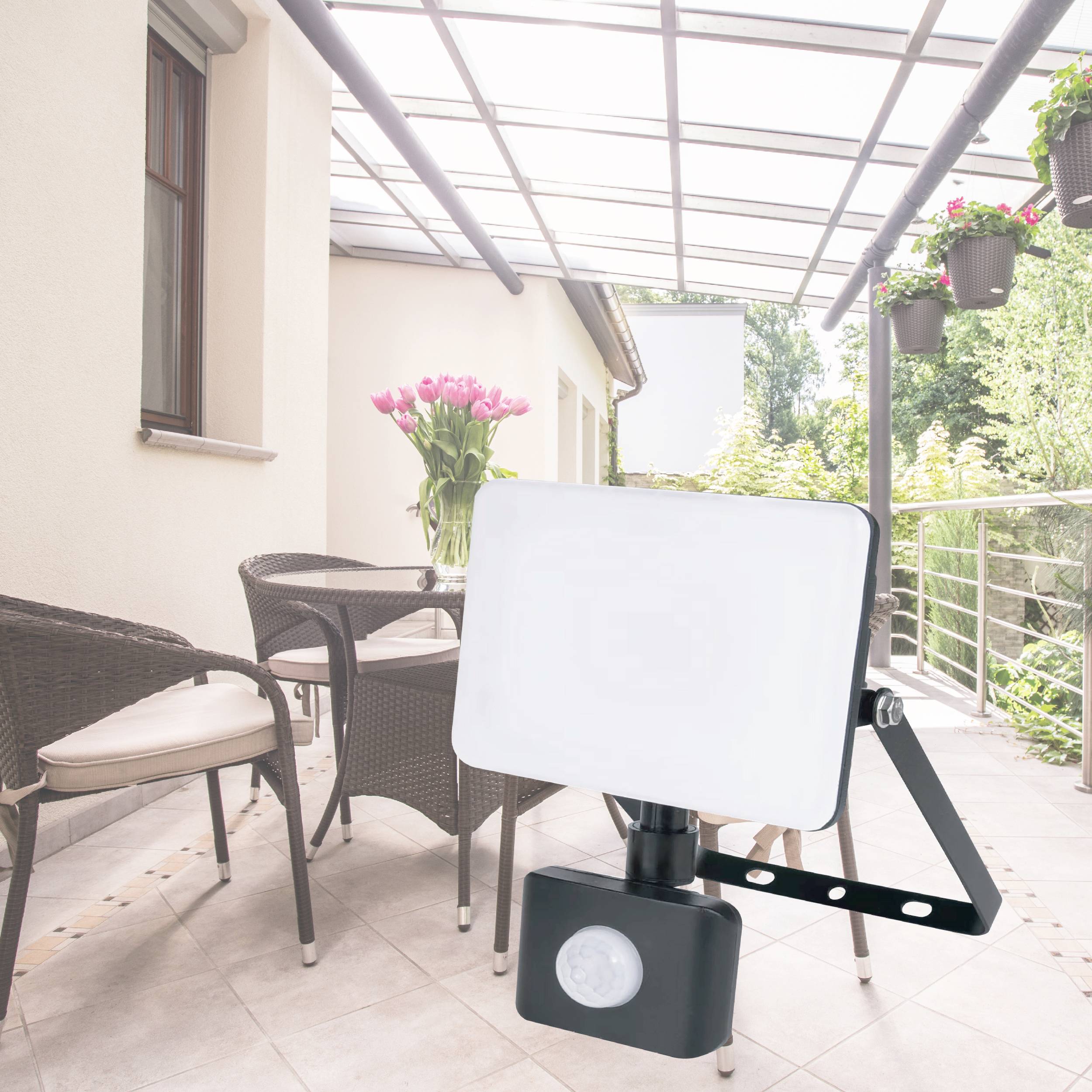 'Balkon mit Sonnenschutz oben, Blumen in Töpfen an Geländer, Stühle und ein Flutlichtstrahler mit Bewegungsmelder auf Fliesenboden.'