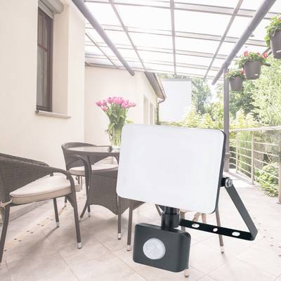 'Balkon mit Sonnenschutz oben, Blumen in Töpfen an Geländer, Stühle und ein Flutlichtstrahler mit Bewegungsmelder auf Fliesenboden.'