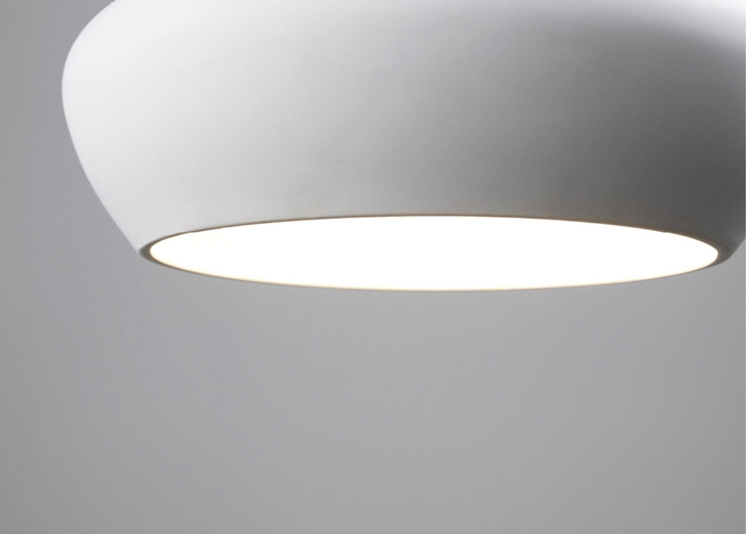 Eine moderne, minimalistische weiße Deckenleuchte strahlt ein sanftes, gleichmäßiges Licht in einem neutralen Raum aus.