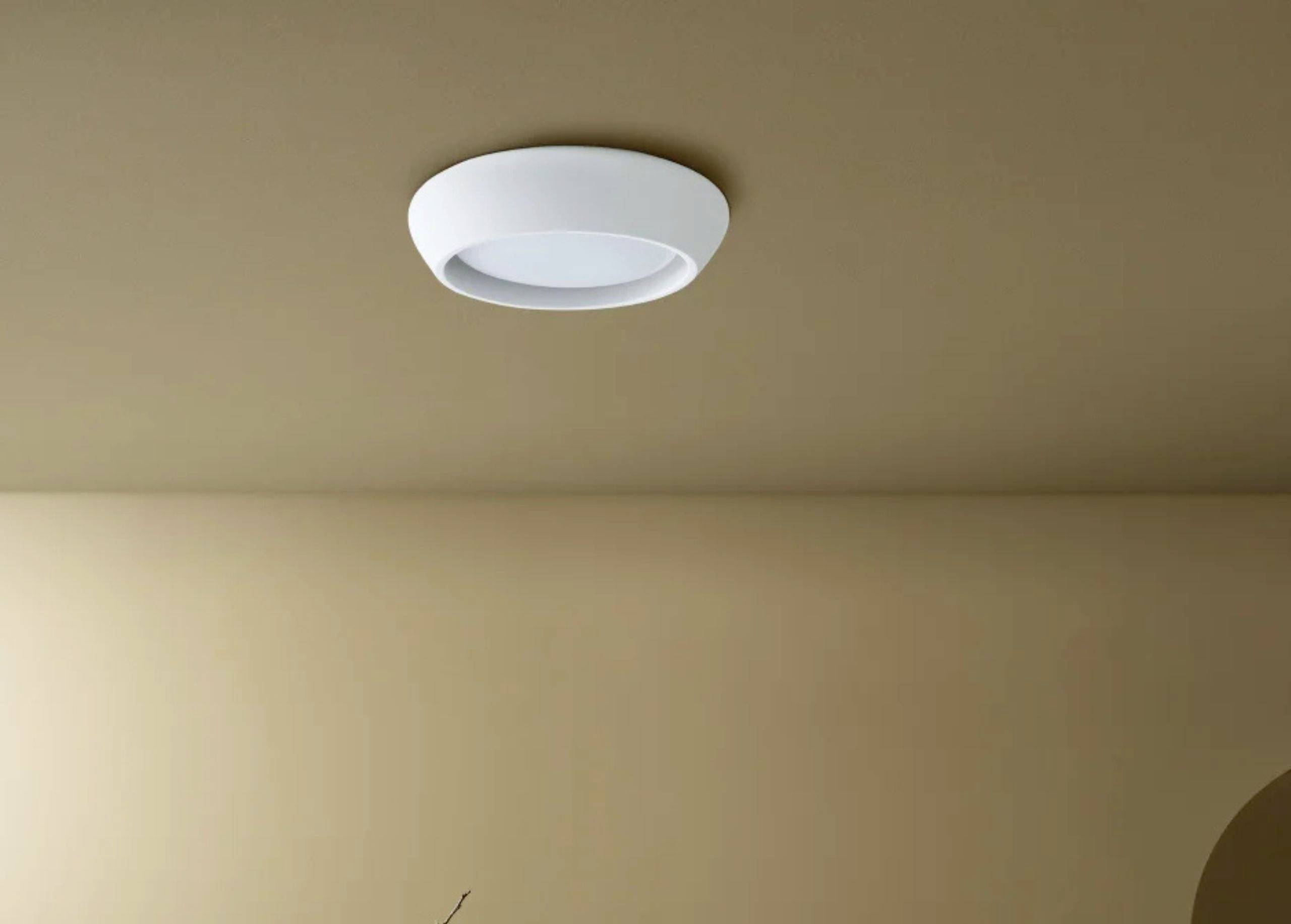 An der Decke eines Raumes ist eine schlichte, runde, weiße Leuchte montiert. Die Wand und Decke haben einen beige Farbton.