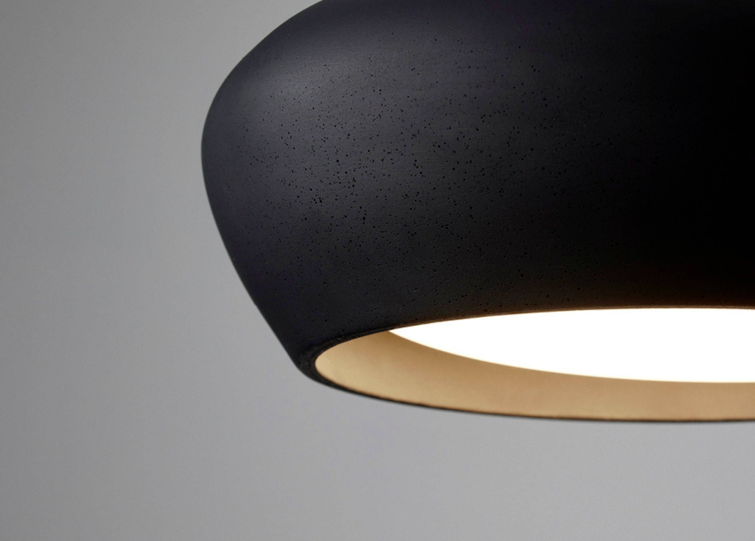 Schwarze, minimalistische Deckenleuchte mit warmem Licht, vor grauem Hintergrund.