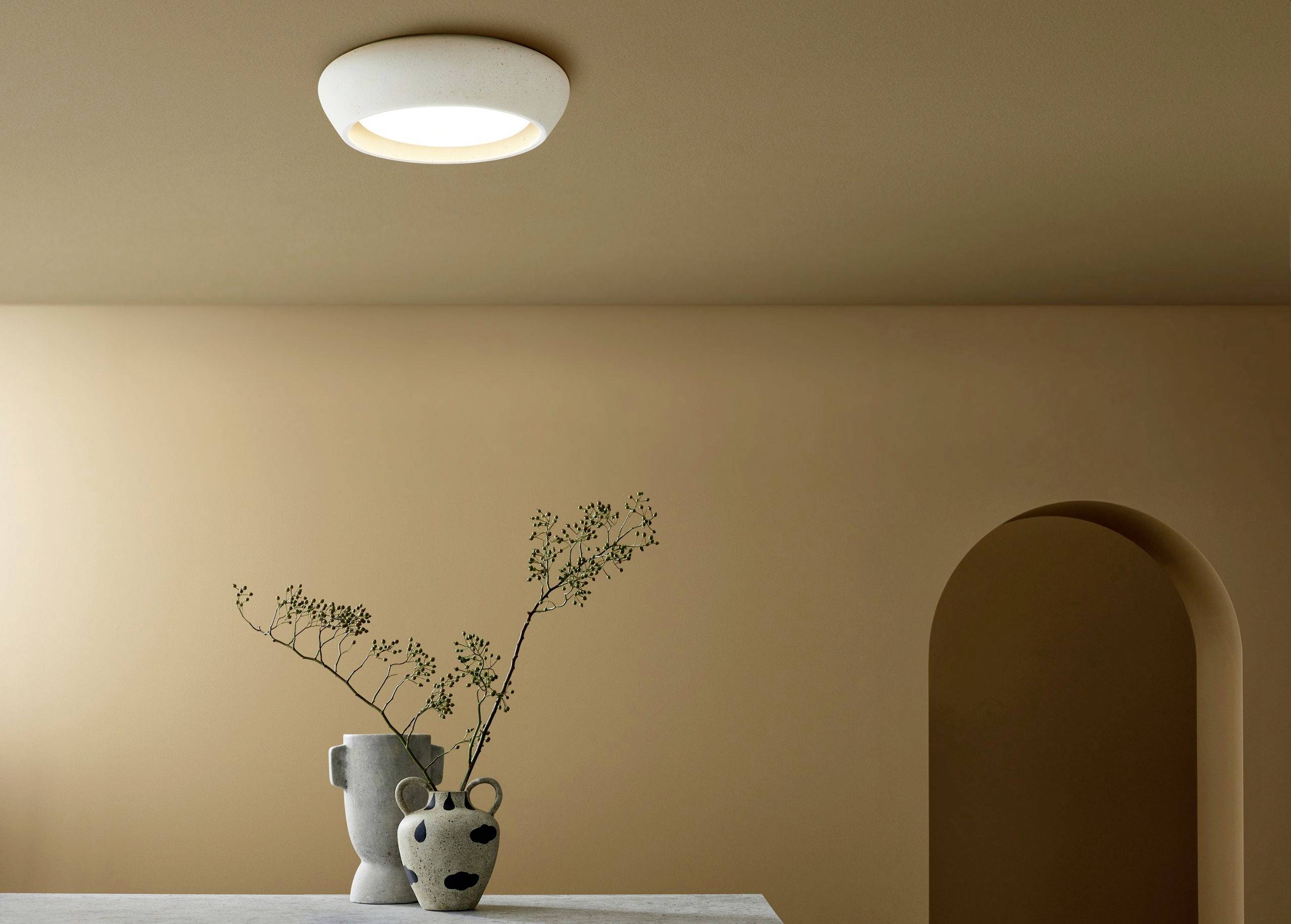 Ein schlicht dekorierter Raum mit einem Tisch, auf dem zwei skulpturale Vasen mit Zweigen stehen. Eine runde Deckenlampe beleuchtet die Szene.