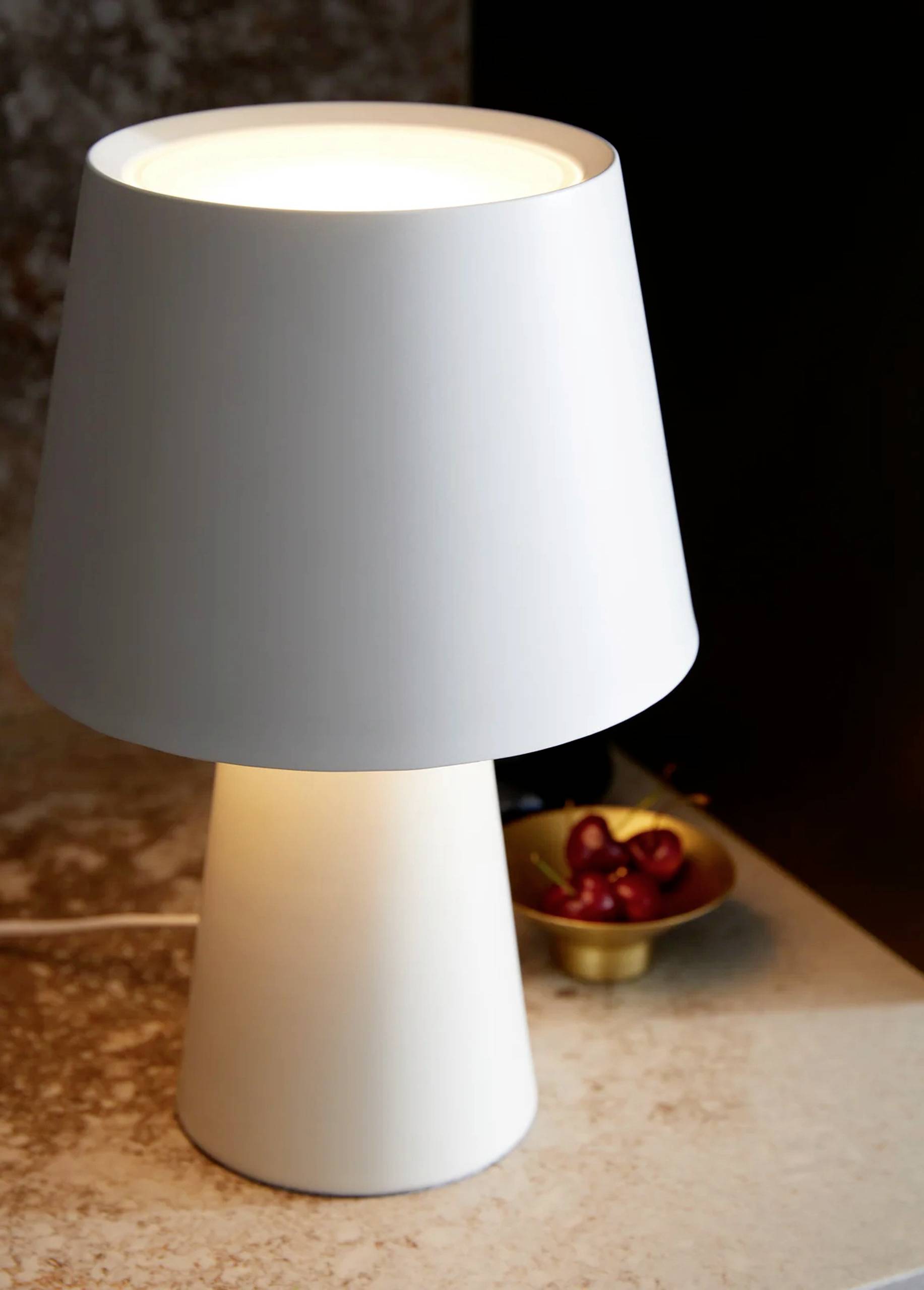 Weiße Tischlampe mit modernem Design auf einem Marmortisch. Daneben steht eine kleine Schale mit roten Beeren.
