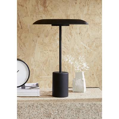 Schwarze Tischlampe mit zylindrischem Sockel auf einem Tisch, daneben ein Stapel Bücher, eine Uhr und eine kleine Pflanze.