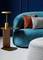 Ein blauer Sessel mit geometrischem Muster, drei bunte Kissen (rot, grau) und ein moderner Holztisch mit goldener Lampe daneben.