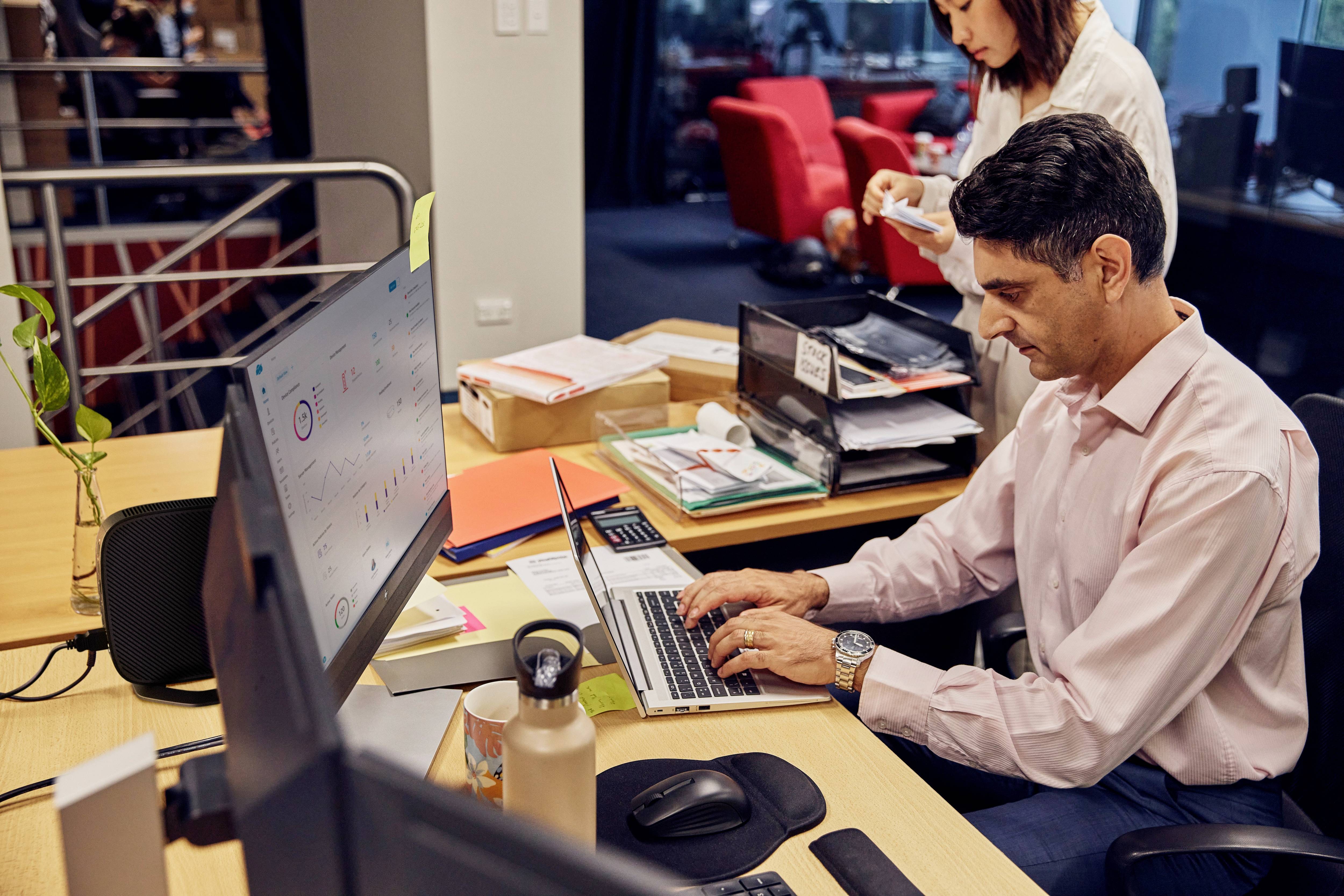 Ein Mann sitzt an einem Schreibtisch und arbeitet am Laptop. Im Hintergrund steht eine Frau mit Notizzetteln. Büro-Umgebung.