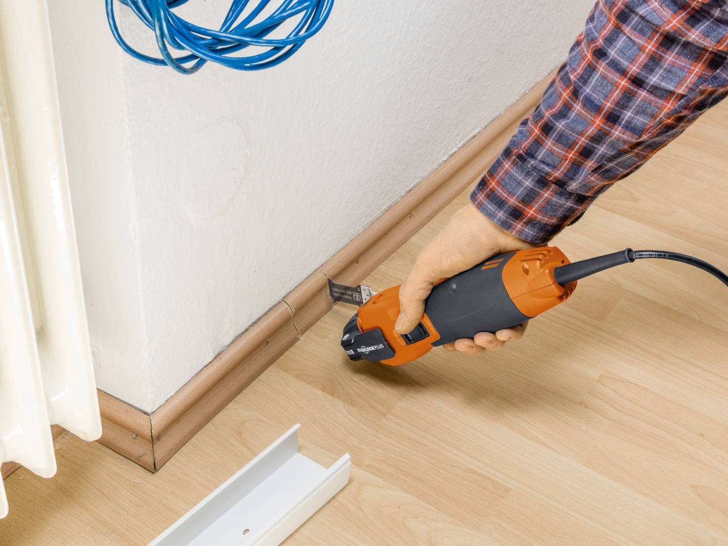 Eine Person schneidet an einer Sockelleiste mit einem elektrischen Werkzeug in der Nähe eines Heizkörpers. Ein blaues Kabel hängt.