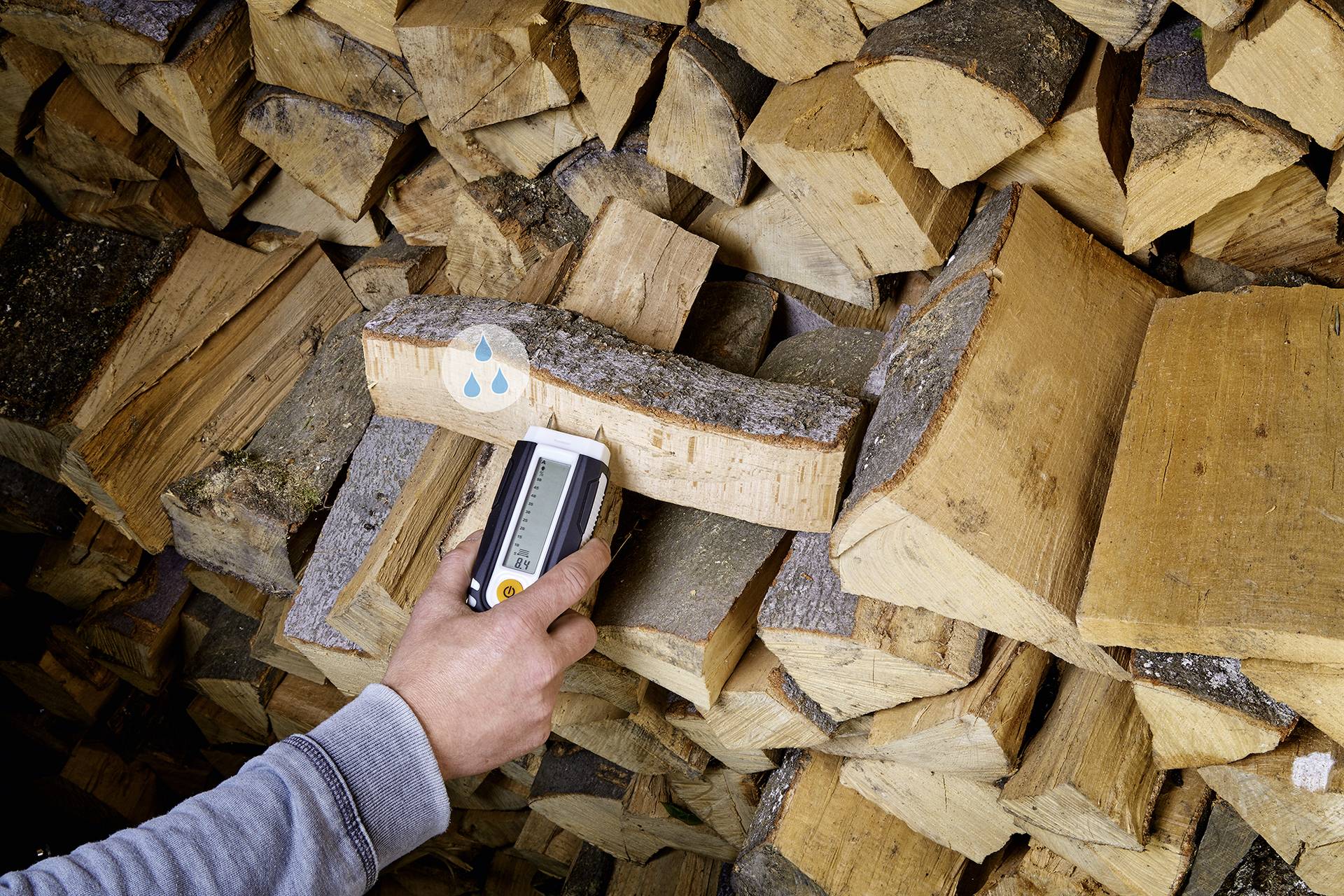 Eine Person misst mit einem Feuchtigkeitsmesser den Wassergehalt eines Holzscheits vor einem Stapel Brennholz.