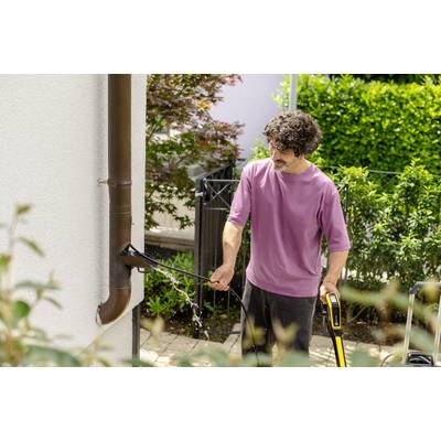 Eine Person in einem lila T-Shirt reinigt mit einem Hochdruckreiniger eine Regenrinne an einem weißen Gebäude. Im Hintergrund sind Büsche.