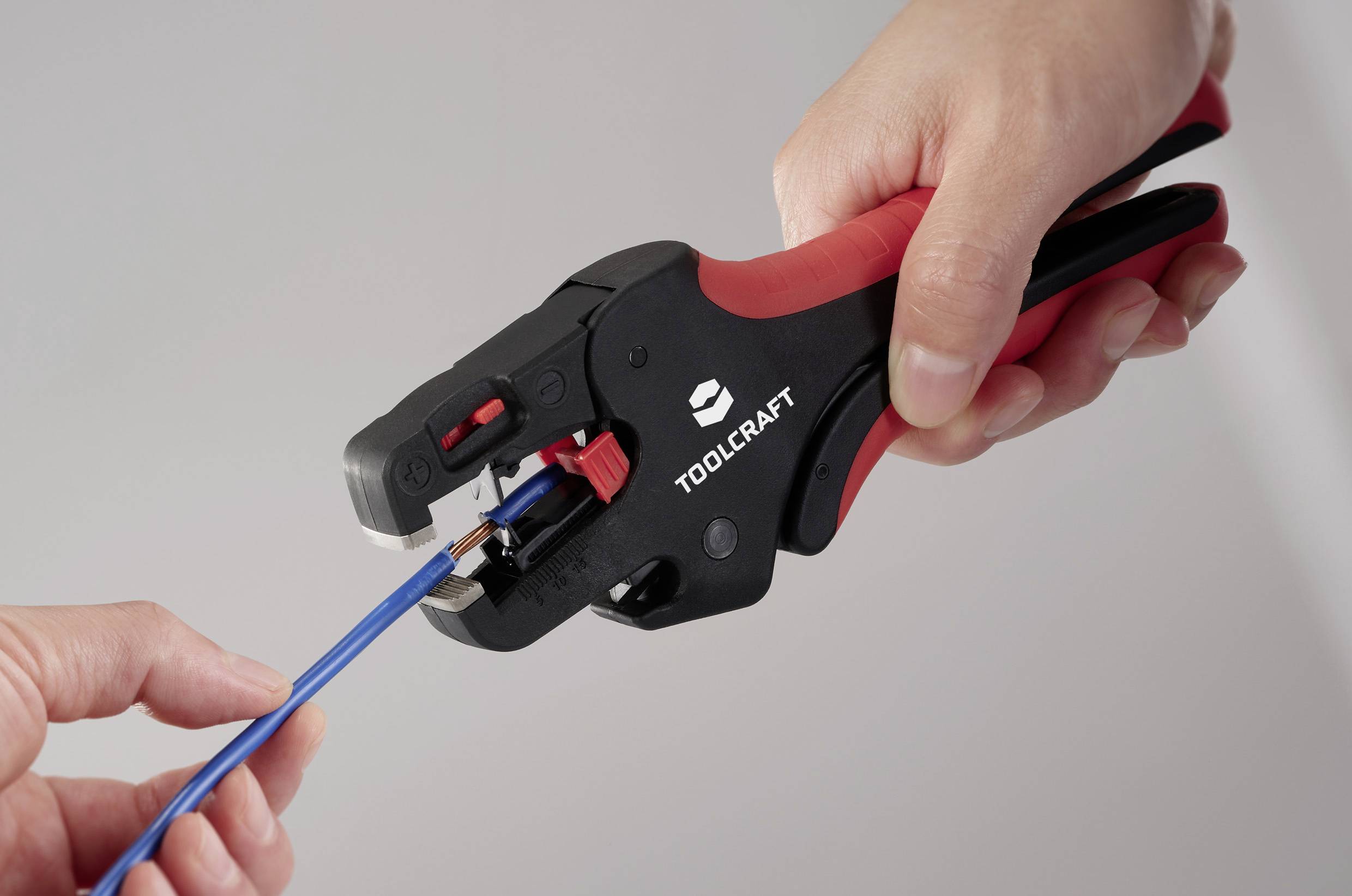Eine Person verwendet ein rot-schwarzes Werkzeug mit der Aufschrift "Toolcraft", um das Ende eines blauen Kabels zu abisolieren.