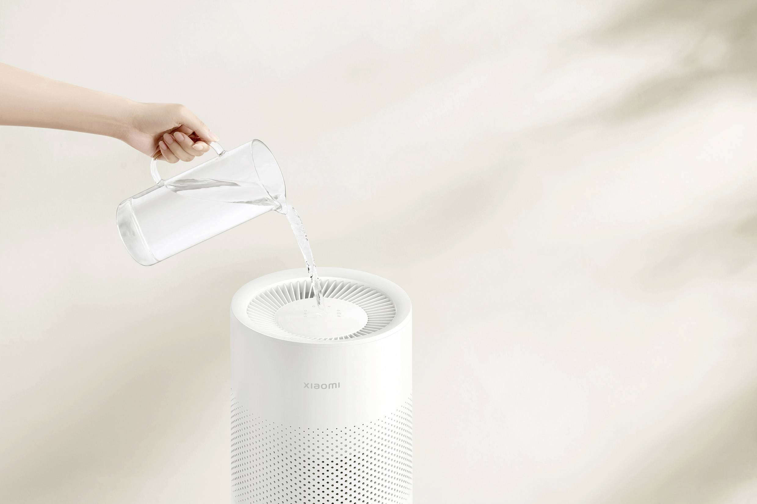 Eine Hand gießt Wasser aus einem Krug in einen Luftreiniger. Der Luftreiniger steht auf einem neutralen Hintergrund.