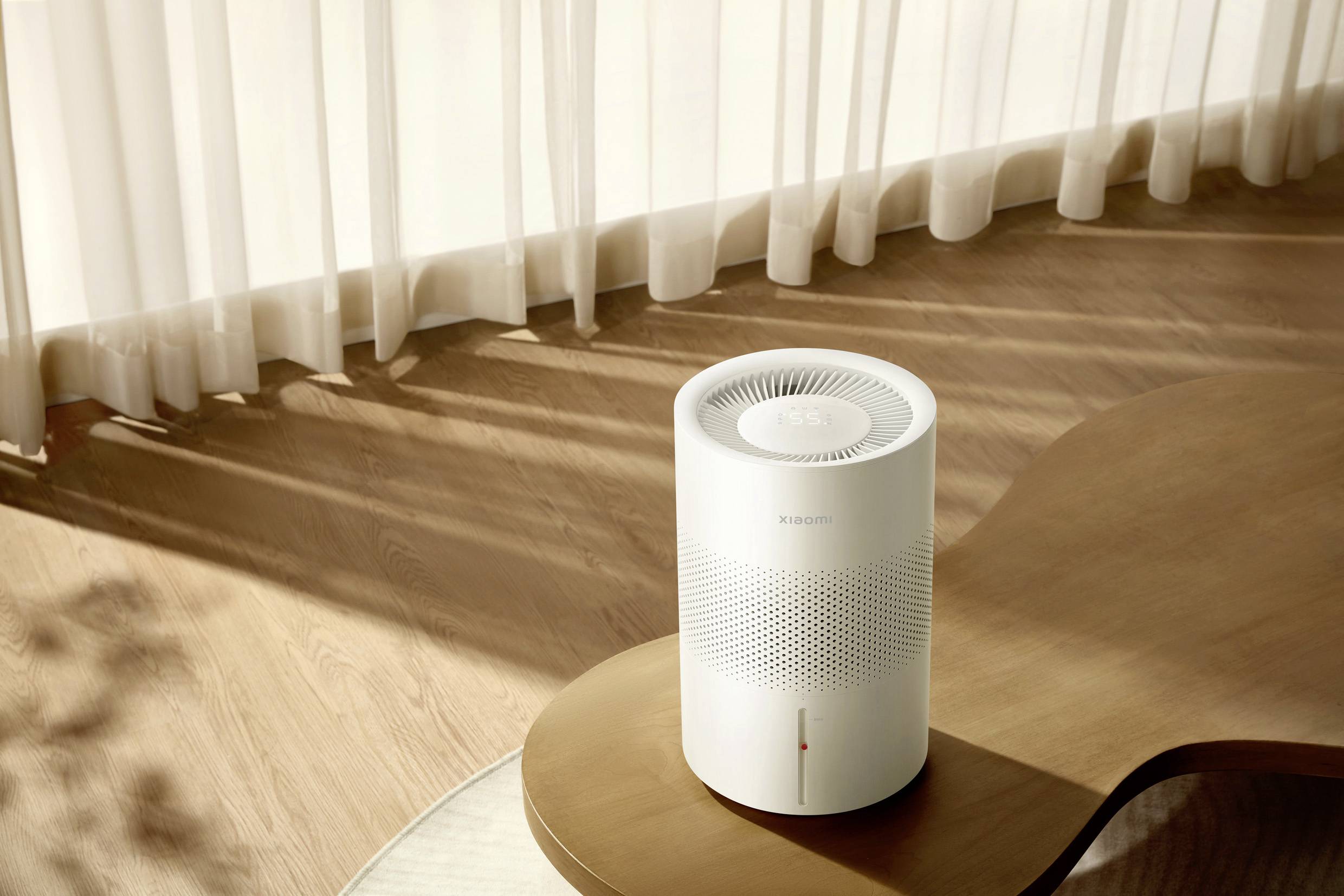 Ein weißer, zylindrischer Luftreiniger auf einem geschwungenen Holztisch. Sonnenlicht fällt durch Vorhänge und erzeugt Schattenmuster.